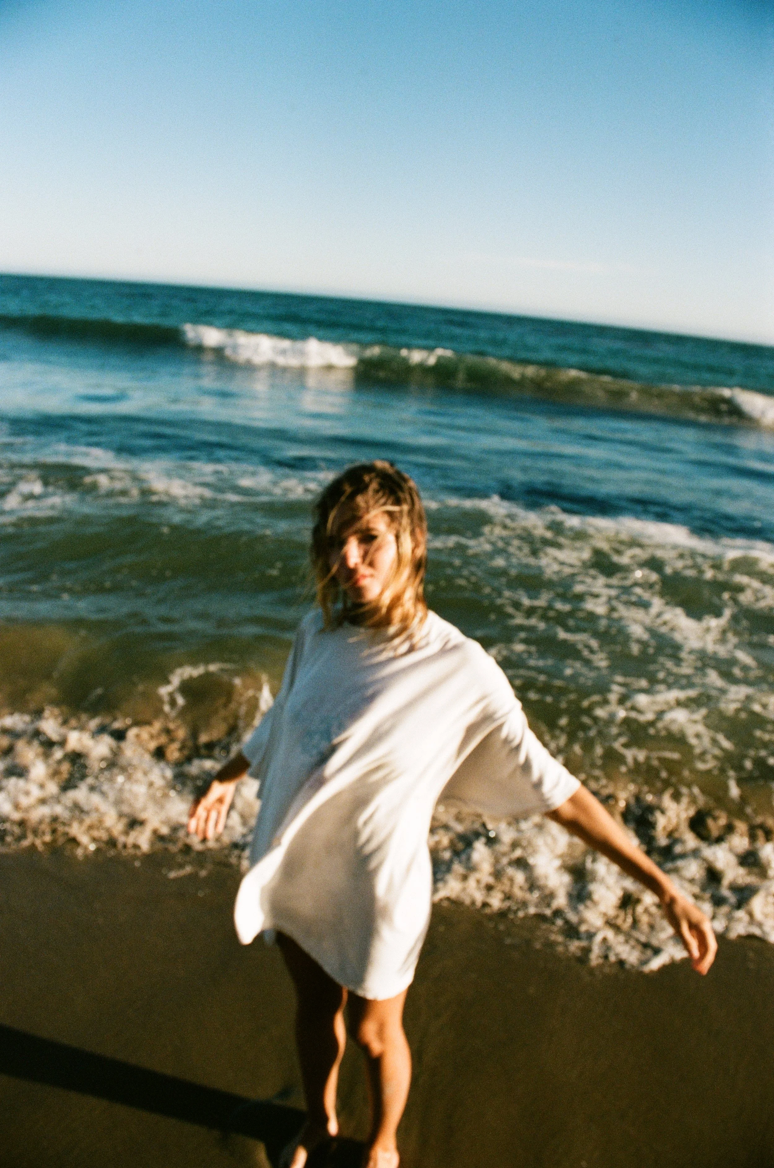 A woman stands on the beach with the ocean and waves behind her, wearing a light-colored dress, with her hair covering her face.