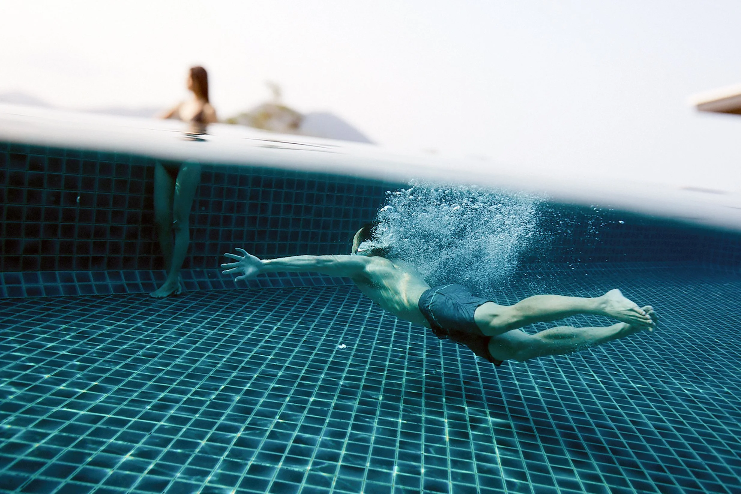 A person diving into a swimming pool underwater with a tiled pool floor, while a person stands outside by the pool's edge.