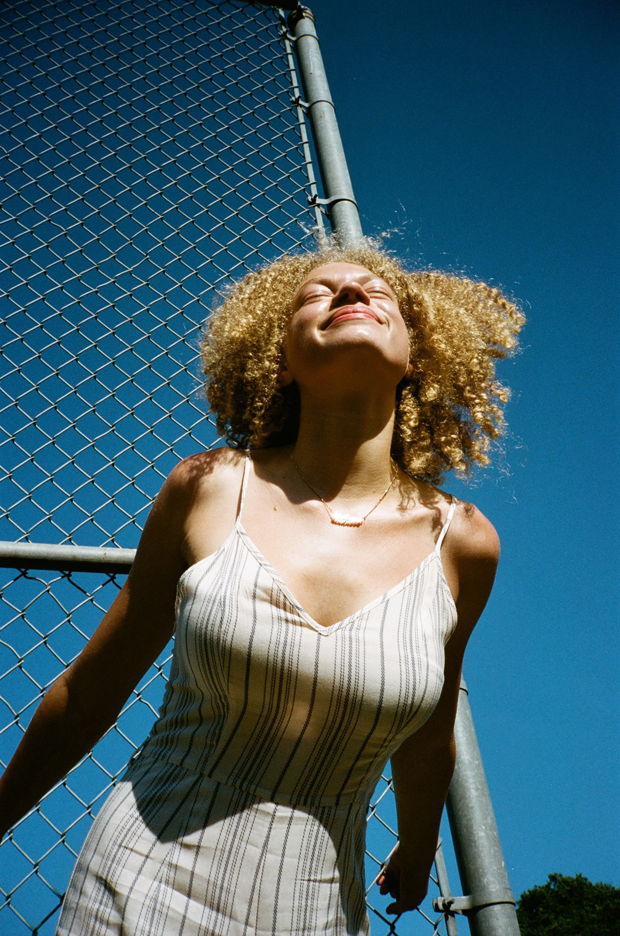 A woman with curly blonde hair smiling with eyes closed, standing in front of a chain-link fence against a bright blue sky.