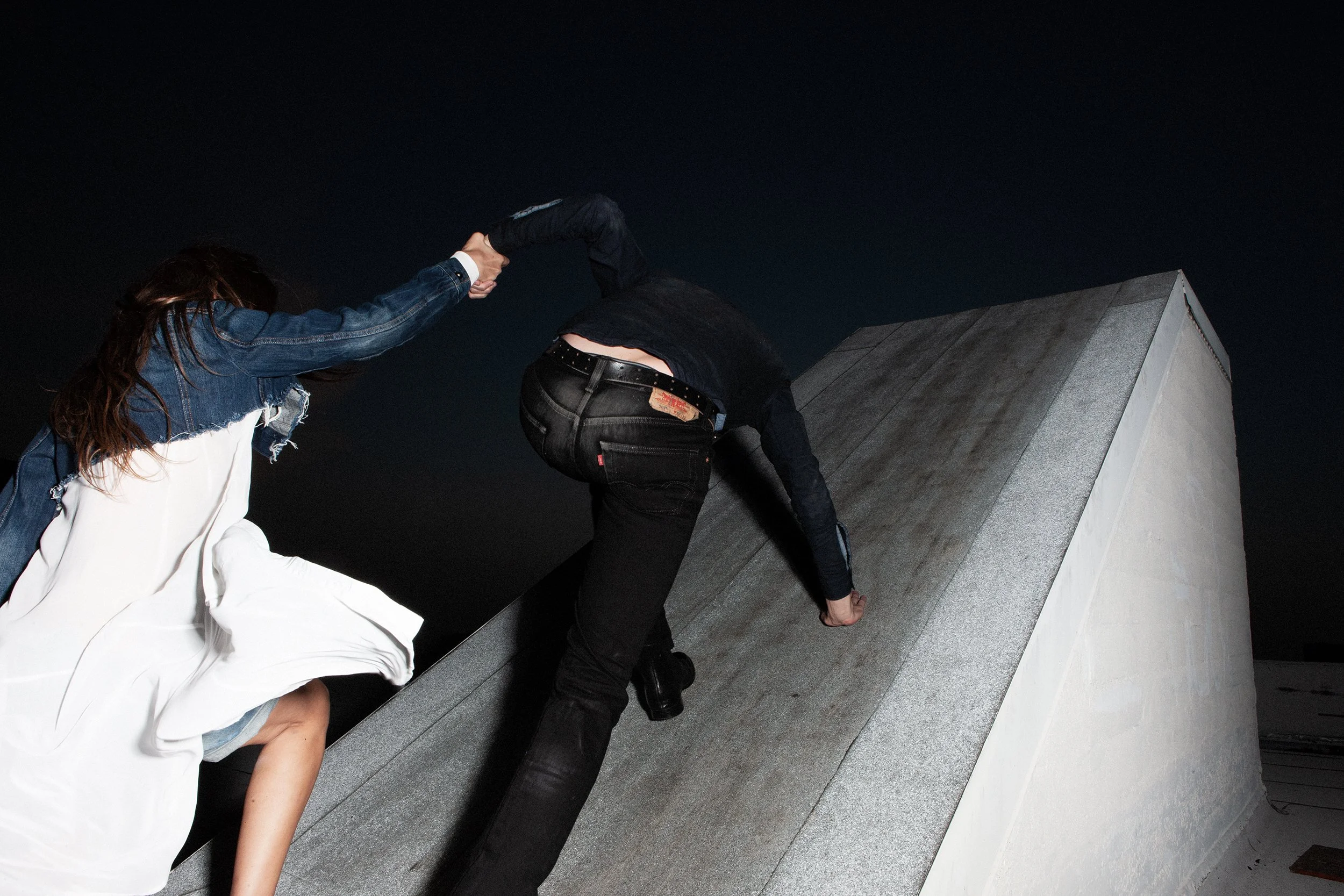 Two people climbing an inclined rooftop at night, one helping the other.