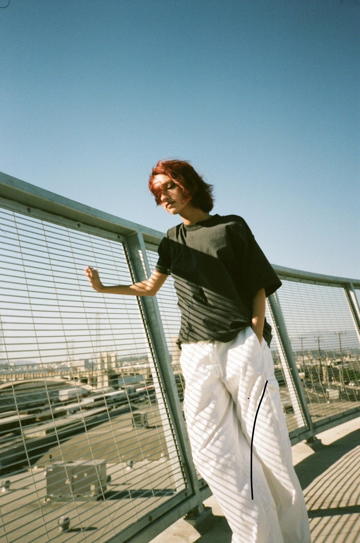 A woman with red hair and sunglasses leaning against a metal railing on a rooftop, with a city skyline in the background and clear blue sky overhead.