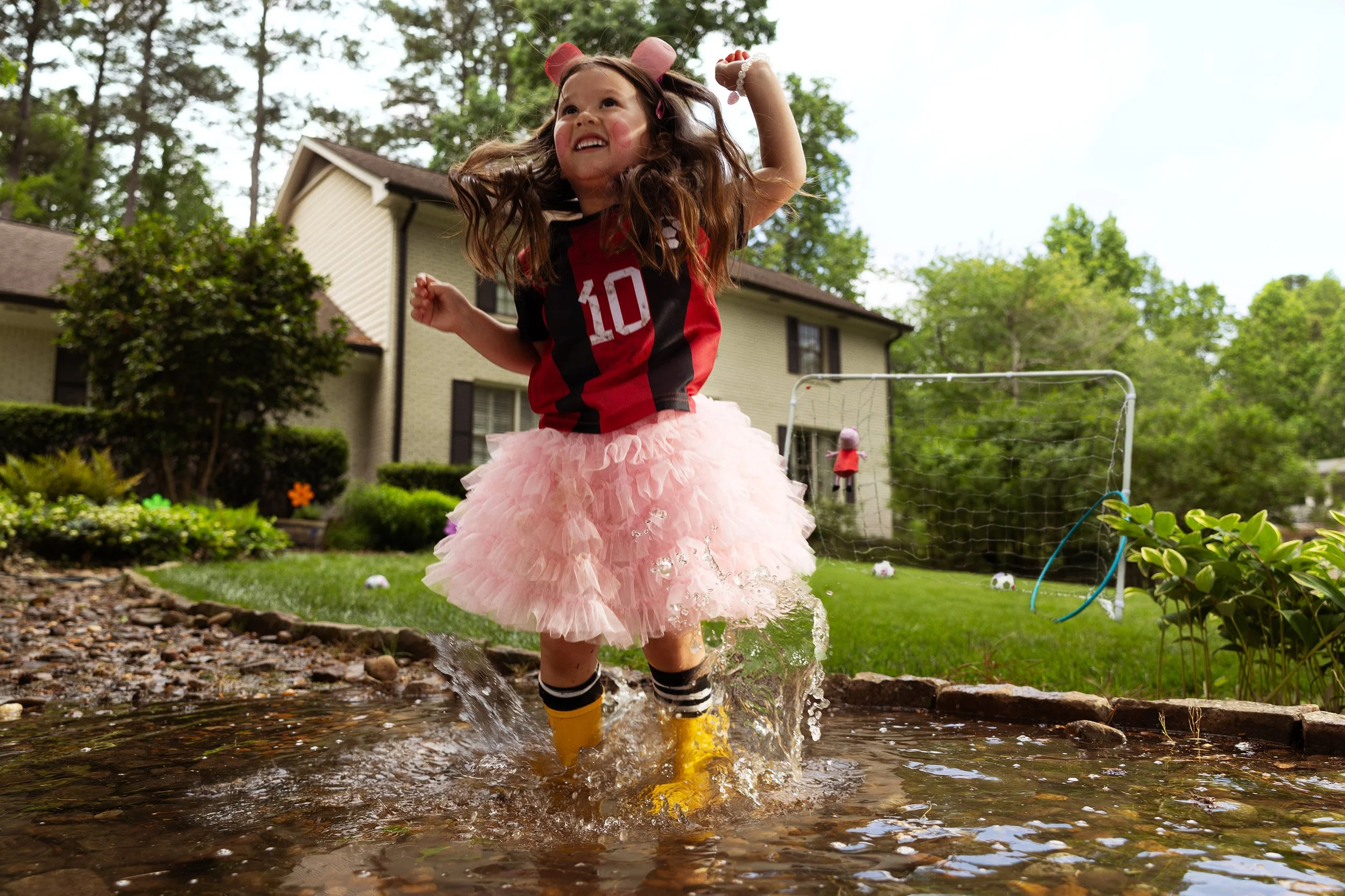 Young girl with long curly hair wearing a red and black sports jersey, a pink tutu skirt, and yellow rain boots playing in a small outdoor pond with a garden, house, and trees in the background.