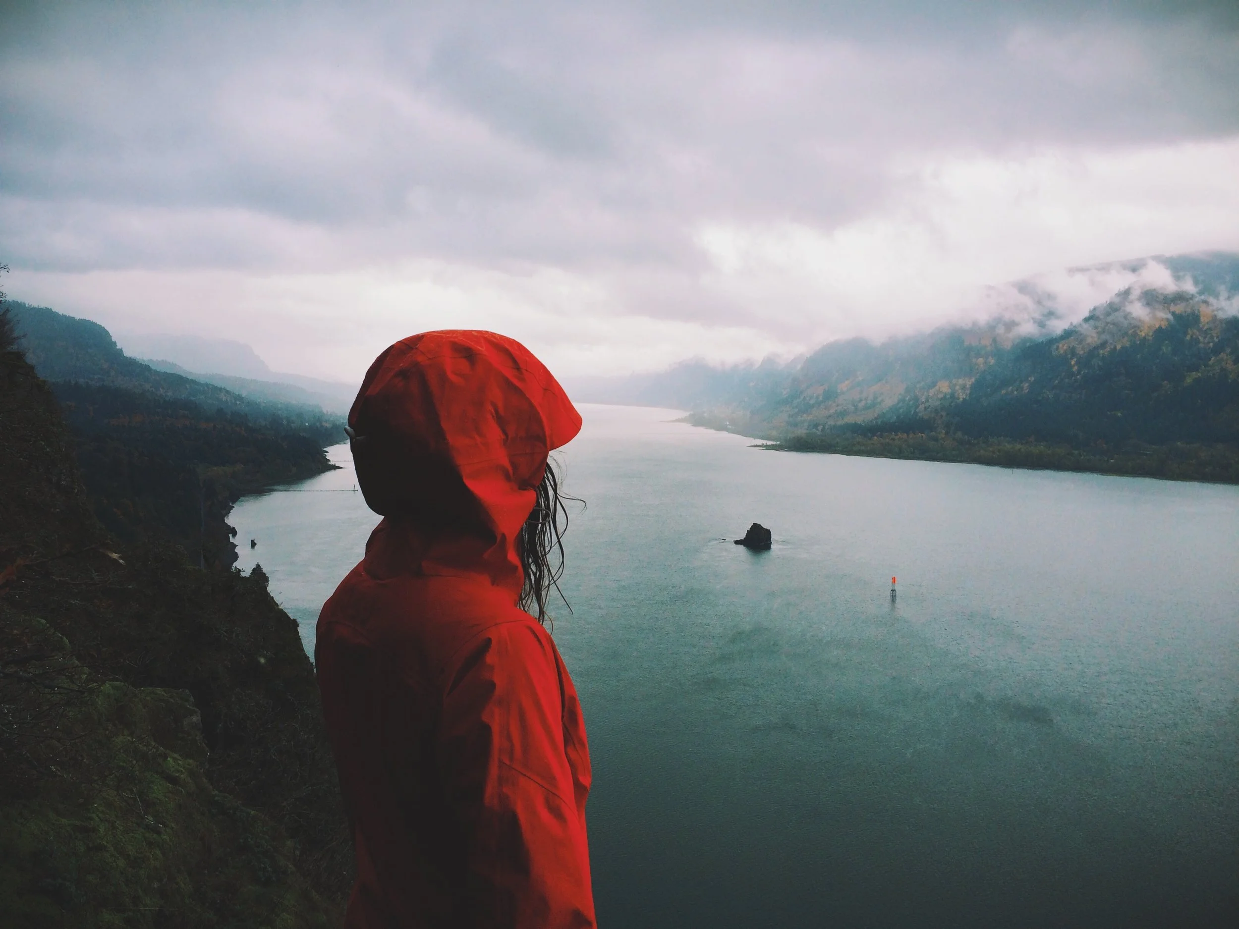Person in a red raincoat looking over a river valley with mountains and clouds in the background.