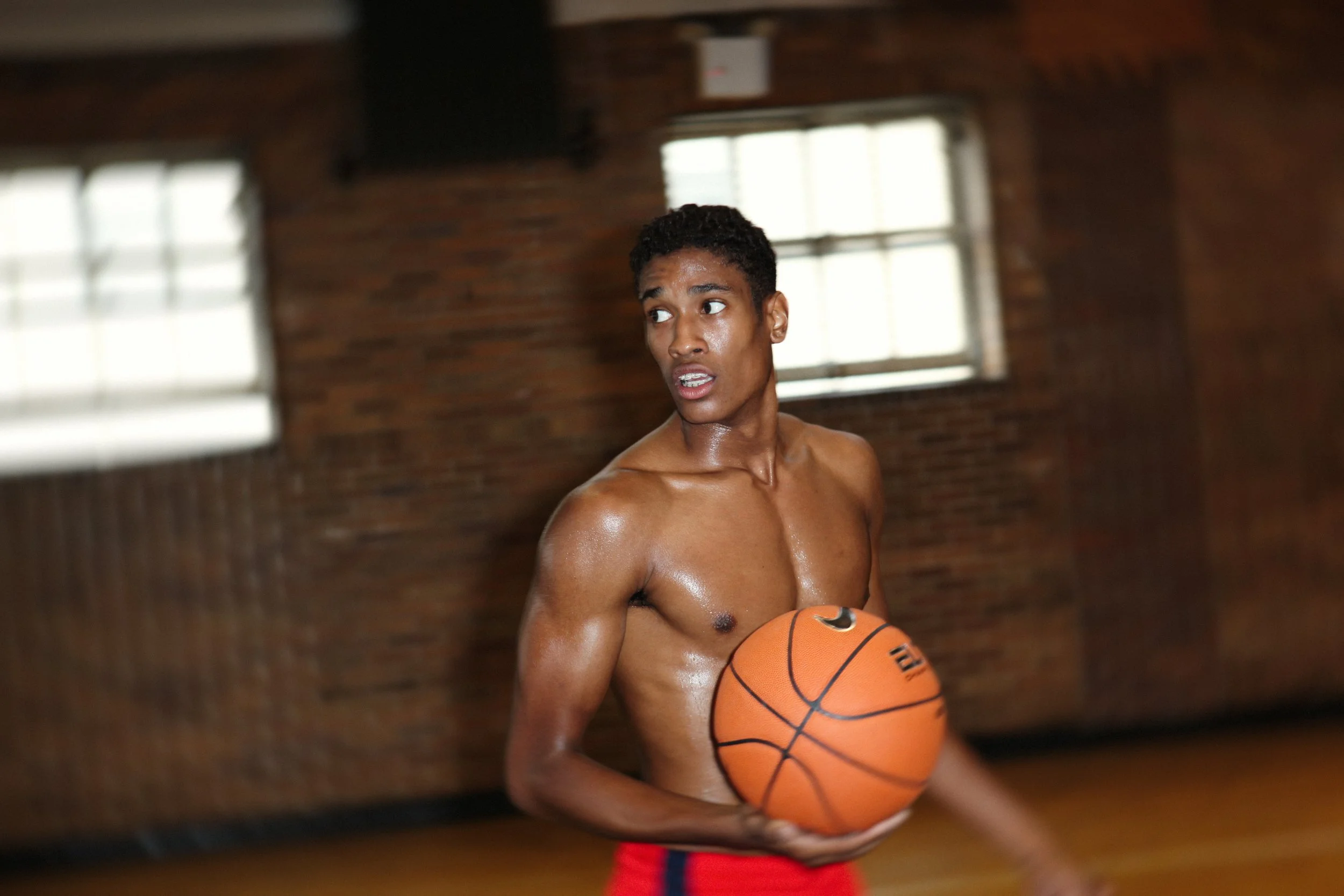 A shirtless young man holding a basketball inside a gym with brick walls and windows.