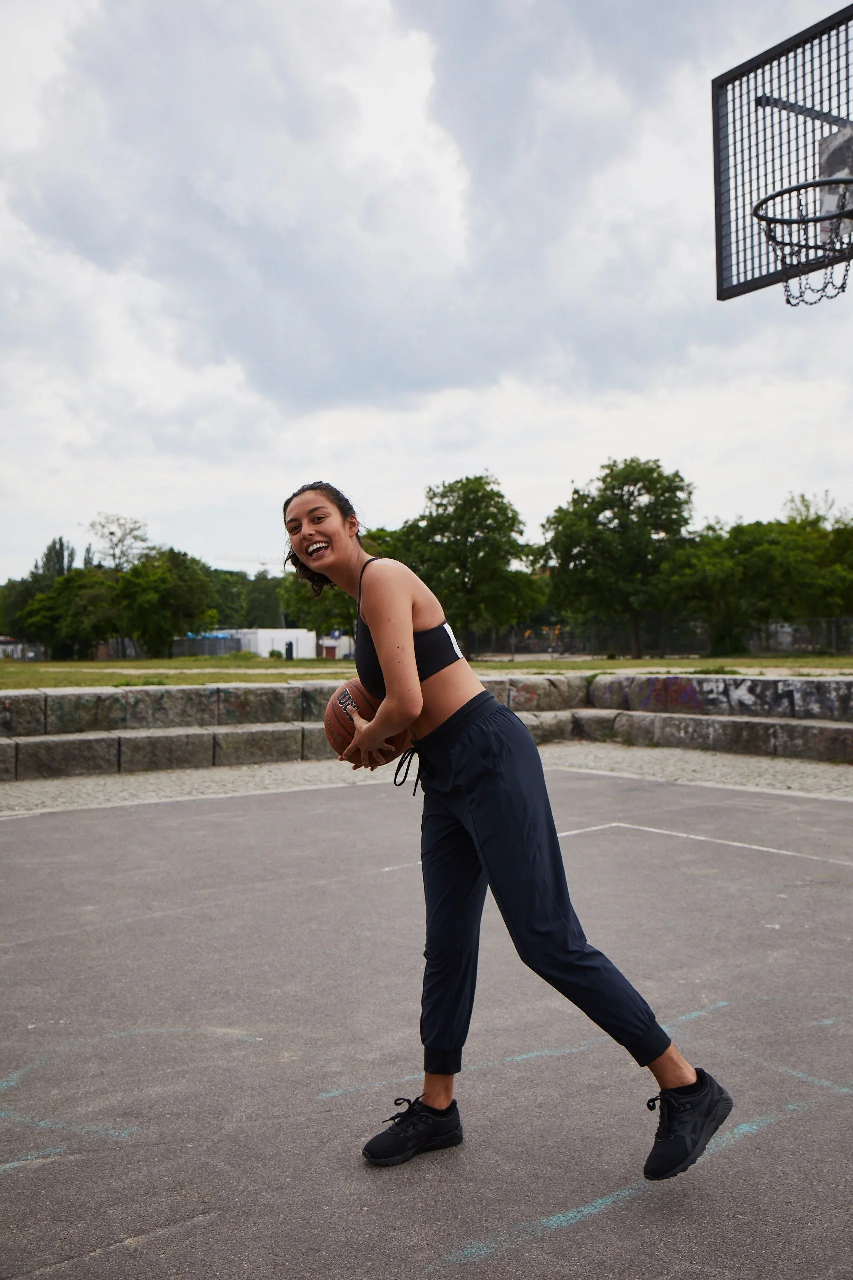 A young woman holding a basketball on an outdoor basketball court, smiling and looking at the camera, under a cloudy sky.