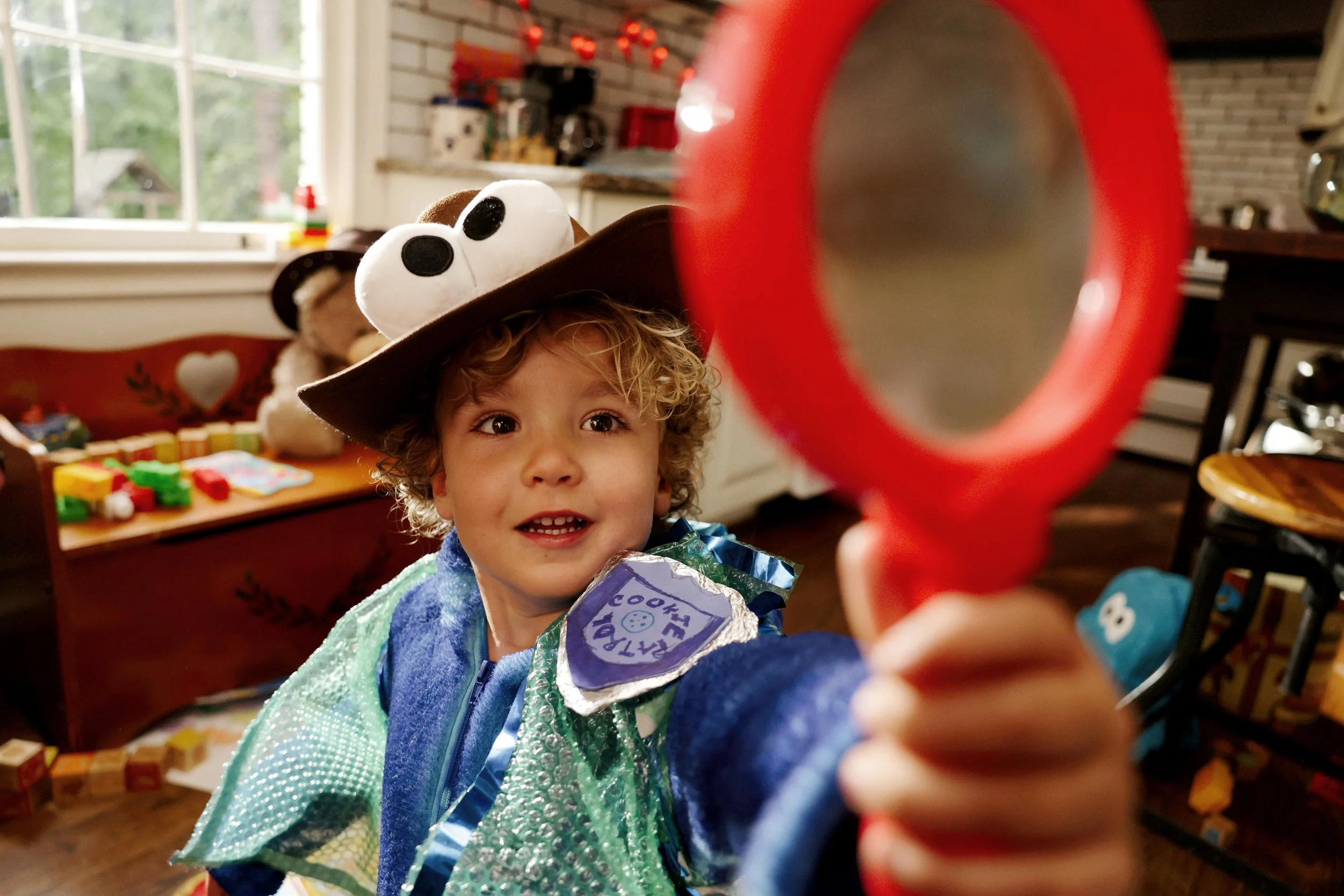 A young child with curly blonde hair wearing a cowboy hat and a shiny green and blue costume, holding a blue and purple toy, and smiling at the camera in a playroom decorated for a celebration.