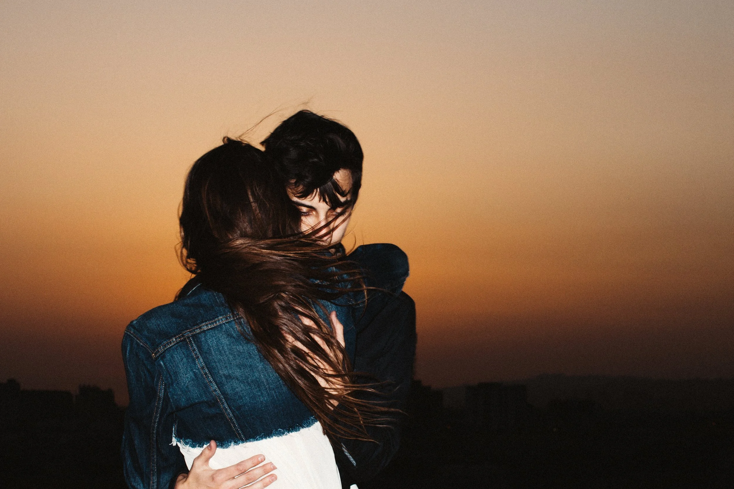 A young couple hugging at sunset or dusk, with the sky displaying warm hues and a silhouette of distant buildings and hills in the background.