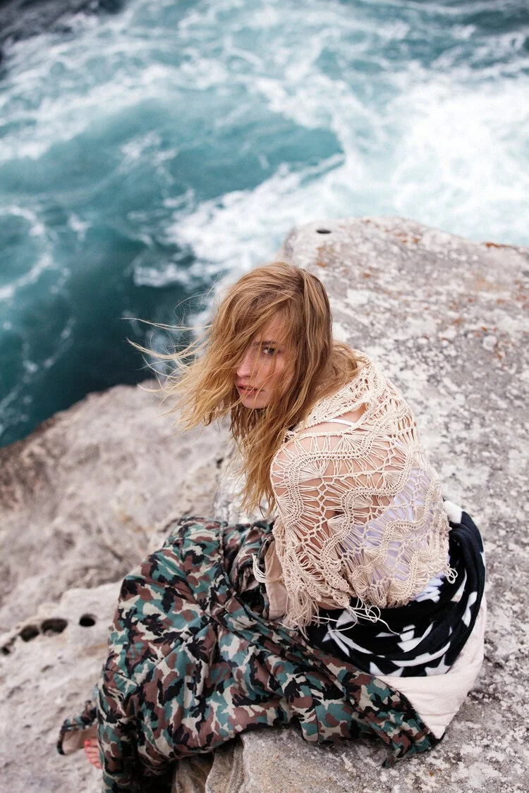 Woman with wet hair sitting on rocky shoreline near ocean, wearing a beige crochet sweater and patterned pants, looking over her shoulder.