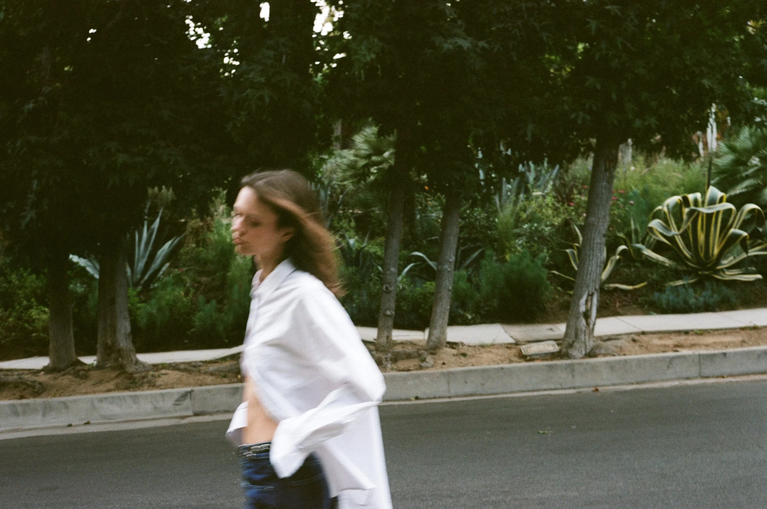 A woman in a white shirt walks along a sidewalk in front of trees and large succulent plants.