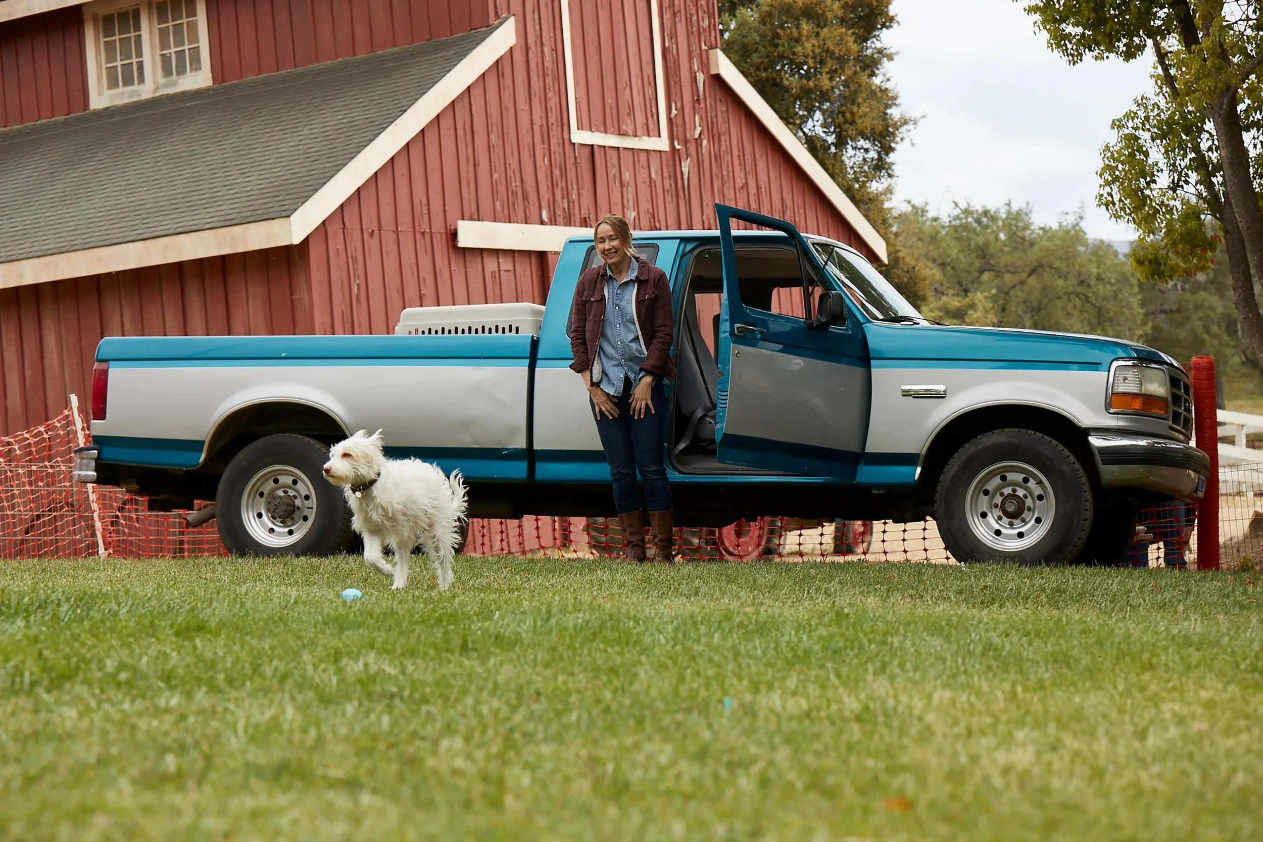 A woman standing beside an open door of a blue and gray pickup truck, with a white dog on a grassy field in front, near a red barn with trees in the background.