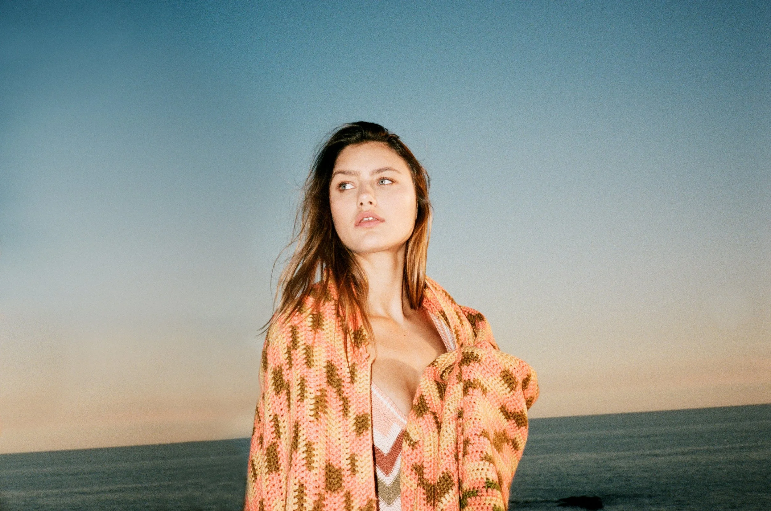 A woman with long brown hair standing by the ocean during sunset, wearing a pink and orange patterned sweater.