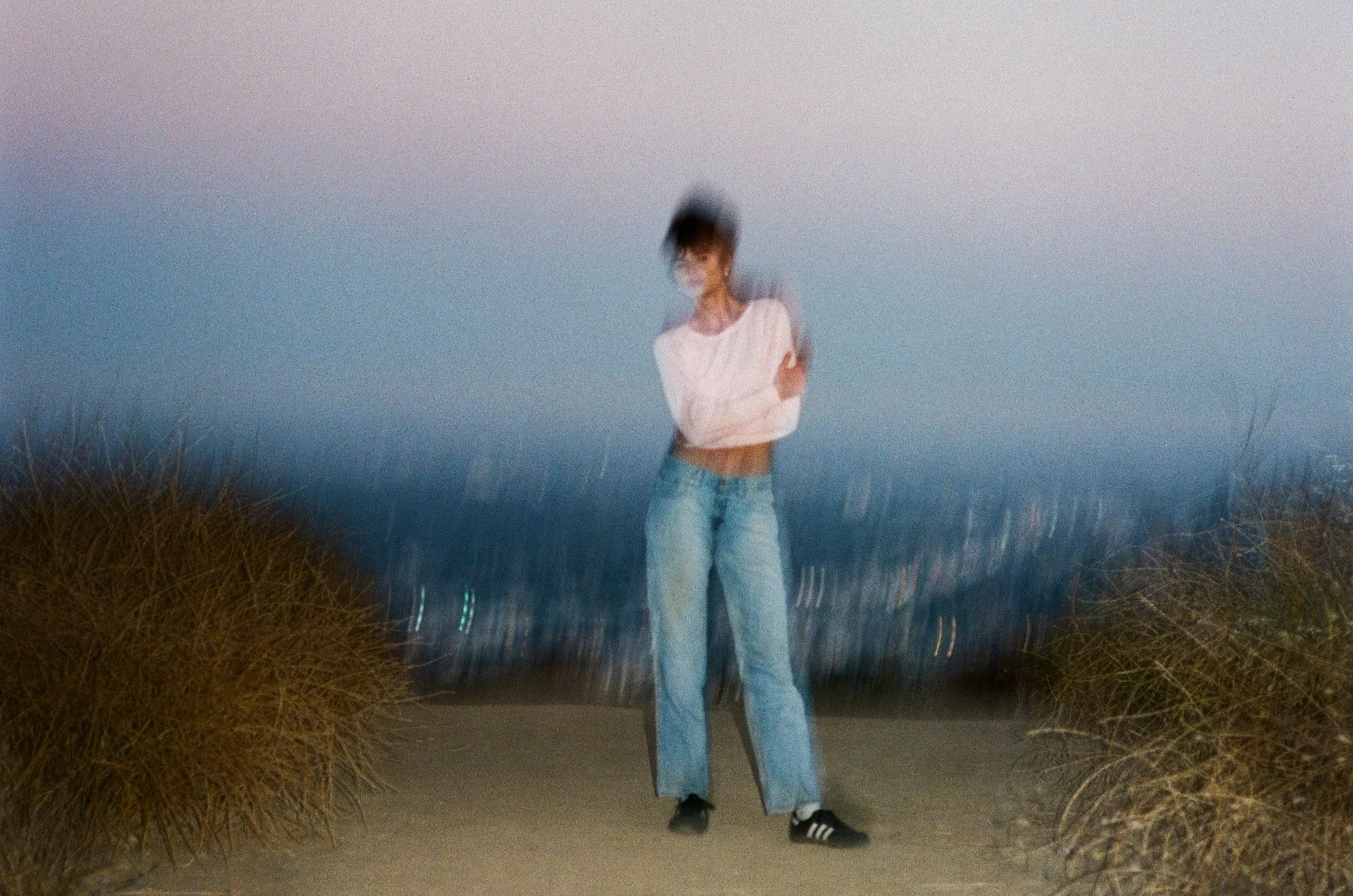 A blurry photo of a person with short hair wearing a light pink long-sleeve shirt, baggy jeans, and black sneakers, standing on a sandy path between two bushes, with a darkening sky in the background.