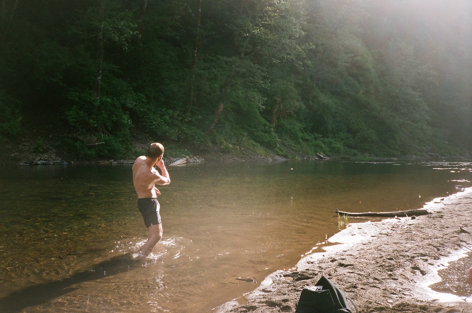 A man standing in a river with a forested shoreline, holding a camera and taking a photo. The sunlight filters through the trees, casting light on the scene.