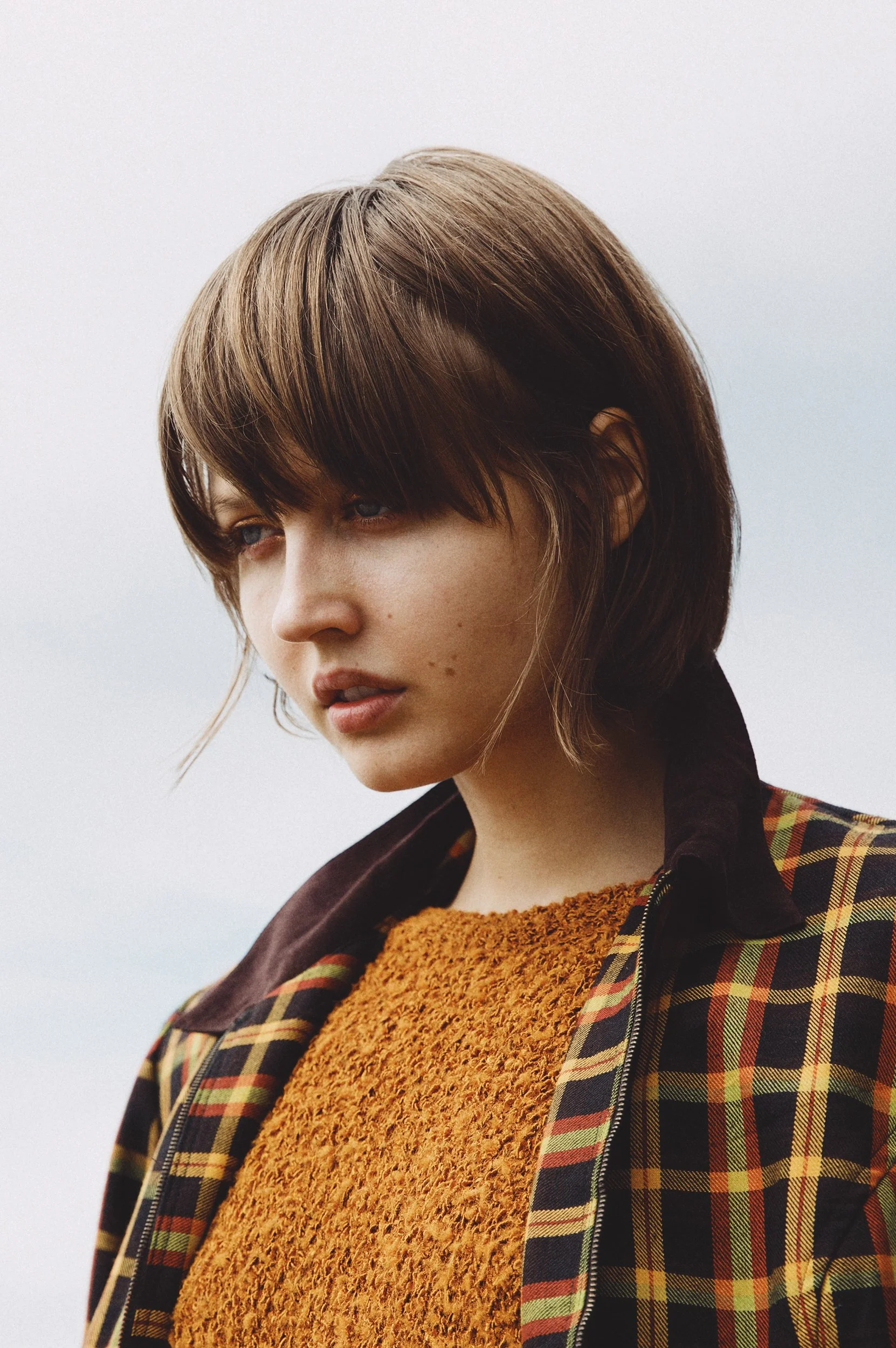 A young woman with short brown hair looking to the side, wearing a plaid jacket over an orange textured sweater, against a cloudy sky background.