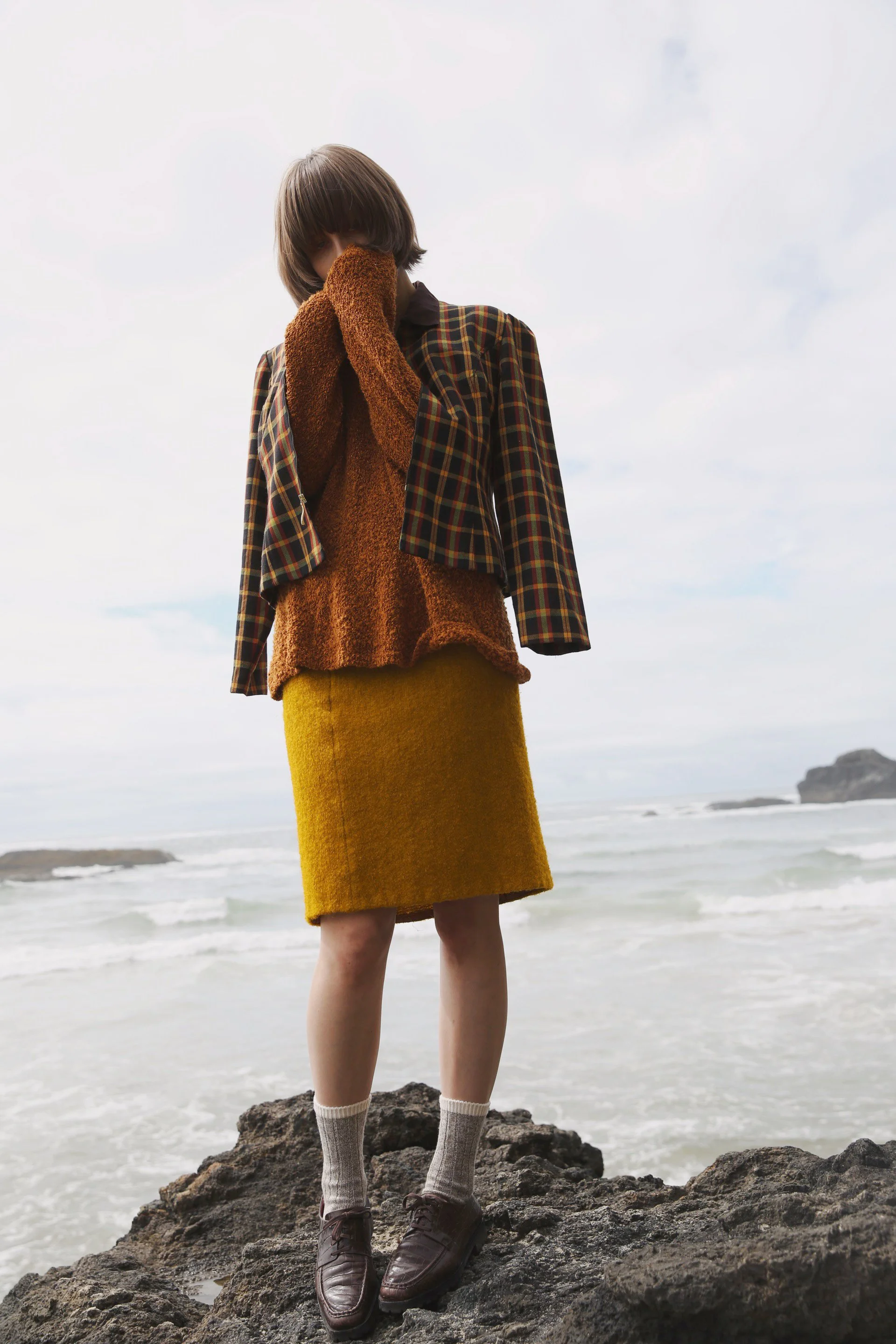 A person wearing a brown sweater, plaid jacket, yellow skirt, and brown shoes with socks standing on rocks at the beach, facing downward with hands covering their face.