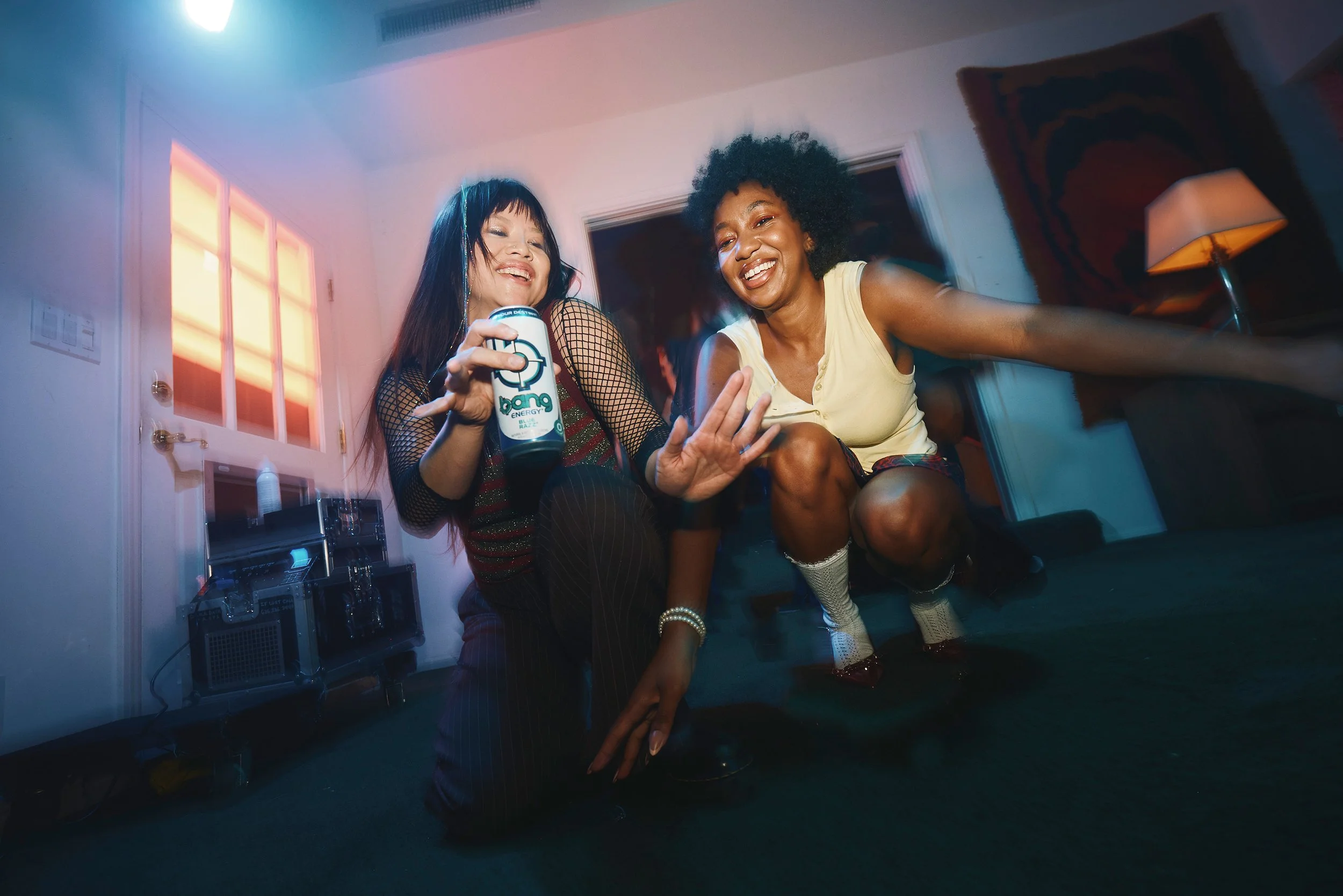 Two women dancing and smiling in a cozy indoor setting, with one holding a can of Bang energy drink, while a lamp and a rug are visible in the background.