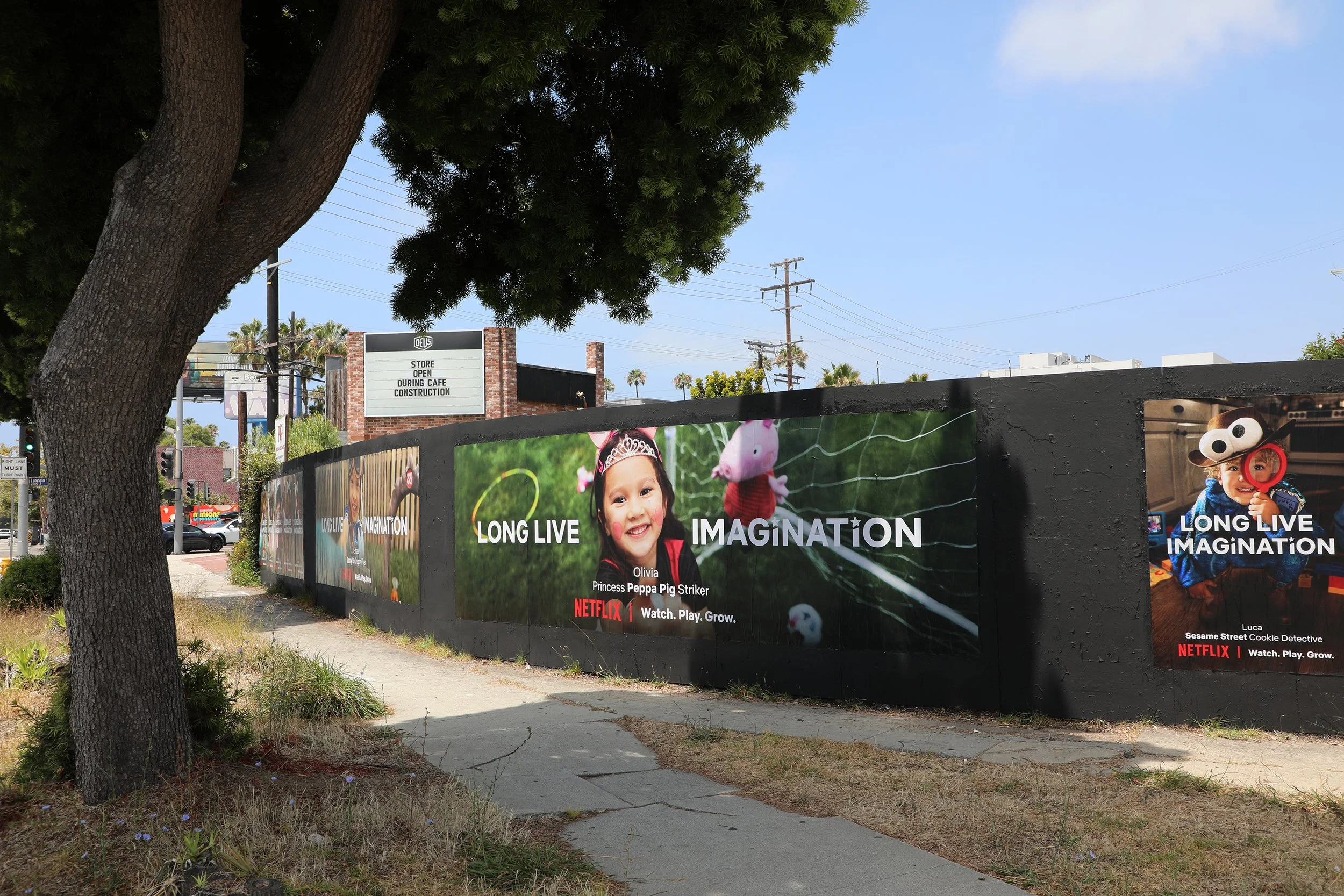 Scene of a sidewalk with large advertising banners featuring children and a pig, trees, utility poles, and a sign indicating a store is open during cafe construction.