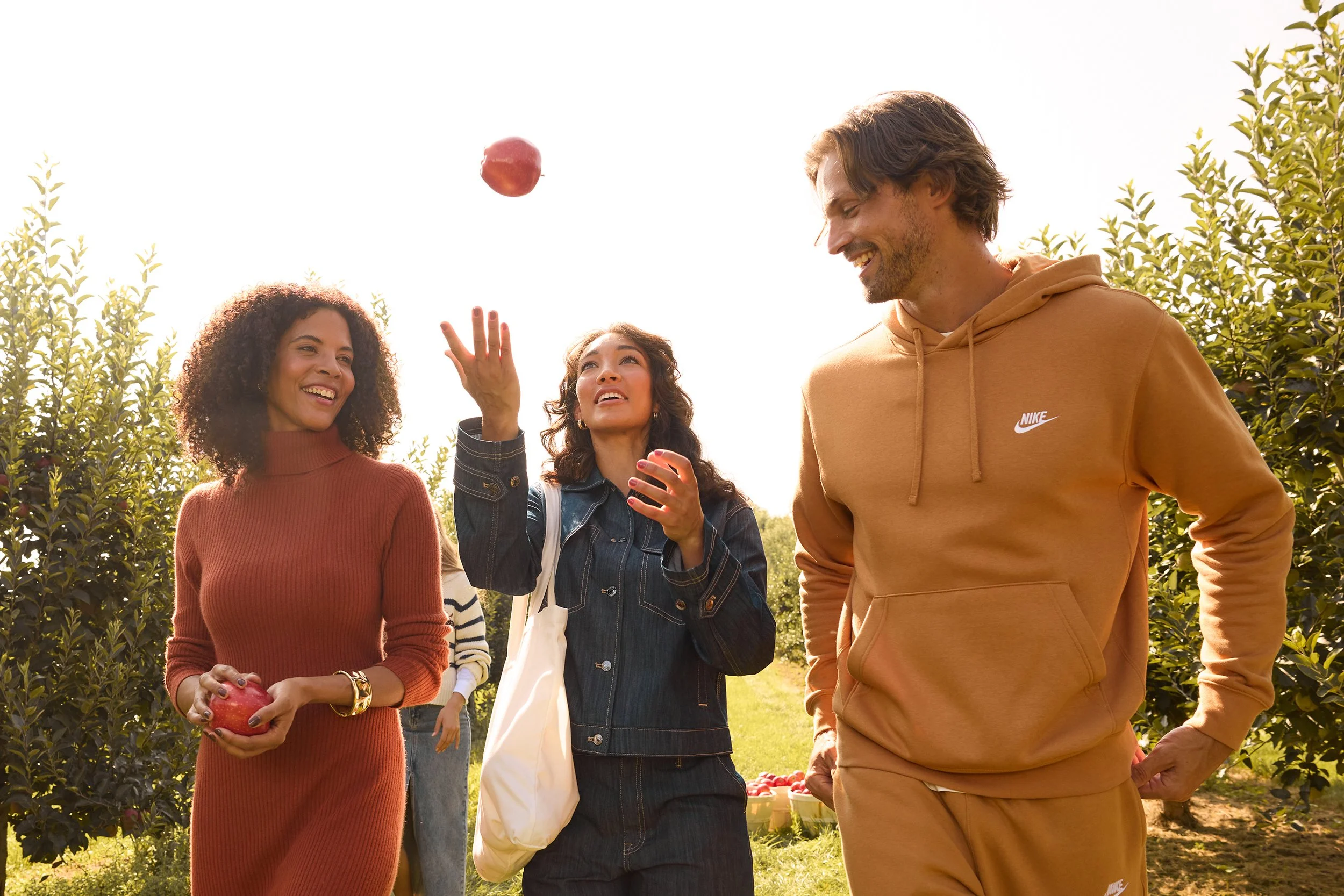 Group of four young adults outdoors during daytime, one tossing an apple in the air, smiling and enjoying the orchard.
