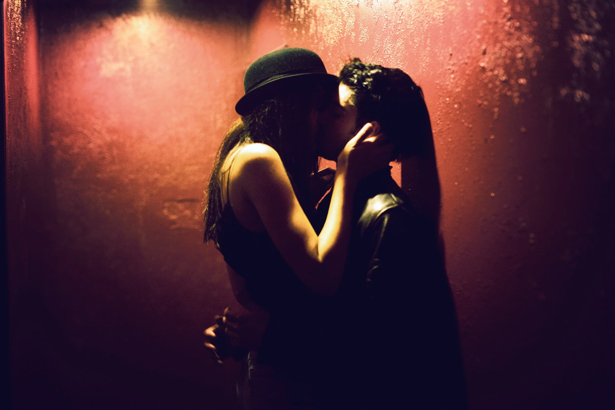 Two people sharing a kiss in a dimly lit setting with a red background, one wearing a hat and the other with short hair, both with dark clothing.