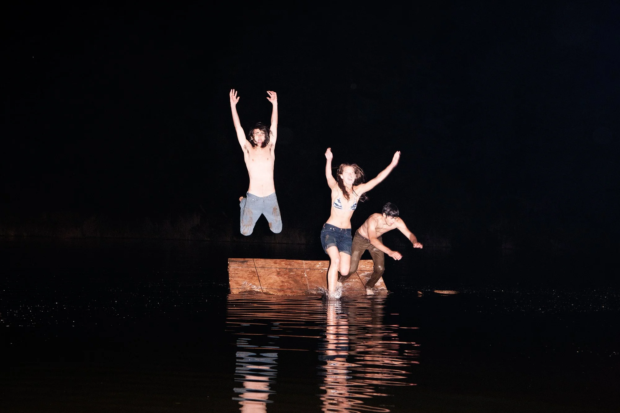 Three people jumping off a wooden platform into dark water at night, with expressions of excitement and surprise.