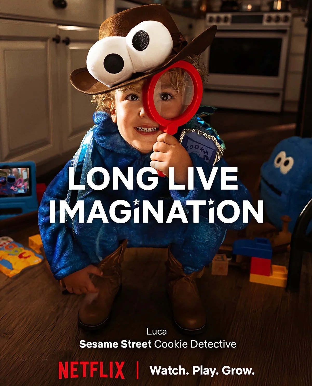 A young boy wearing a blue costume, brown cowboy boots, and a playful hat with large eyes, holding a red magnifying glass. The background includes toys and an oven, and the text overlay reads, 'LONG LIVE IMAGINATION,' with additional details about Lu