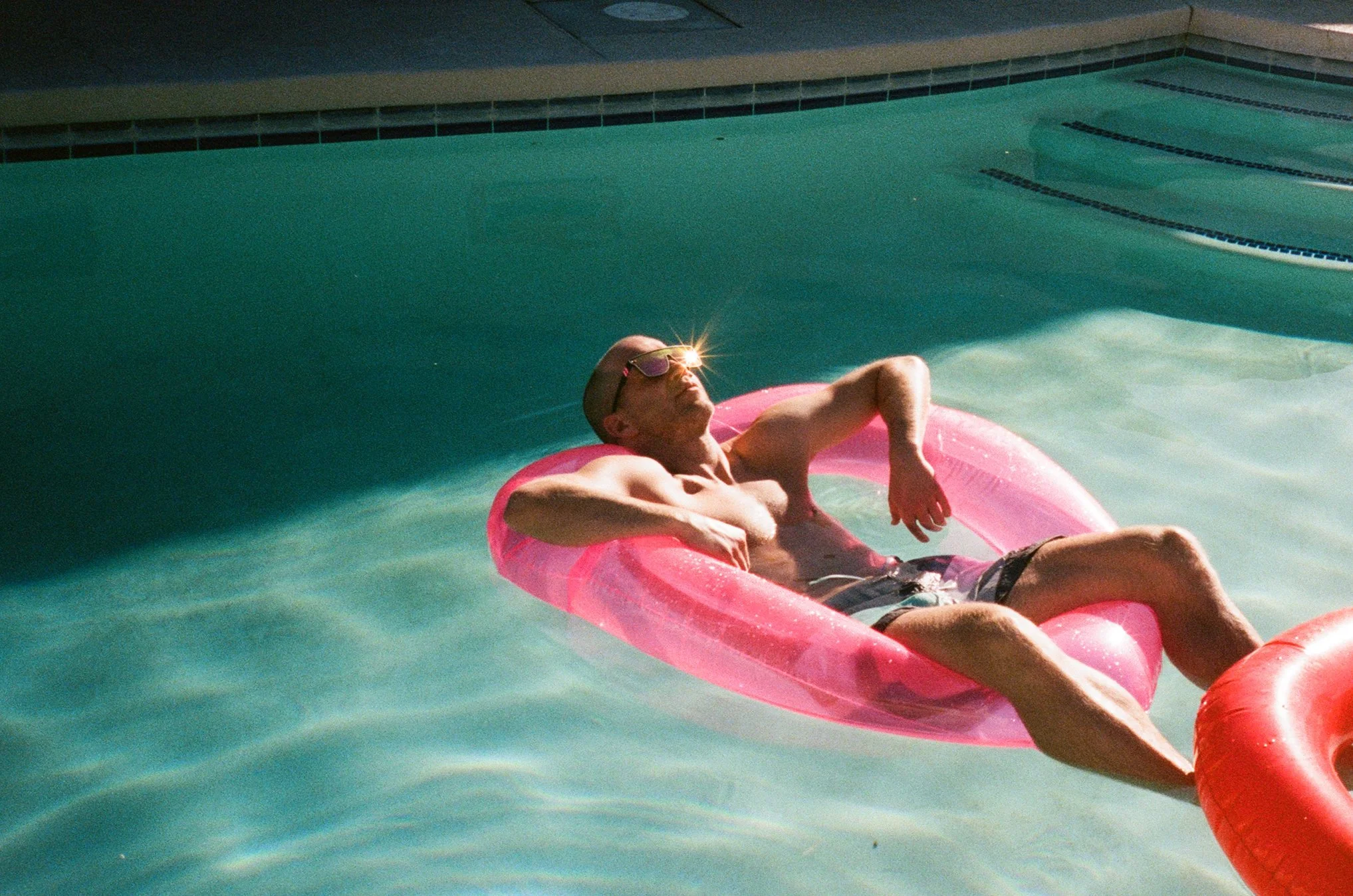 A man relaxing on a pink inflatable float in a swimming pool, wearing sunglasses and swimming trunks.