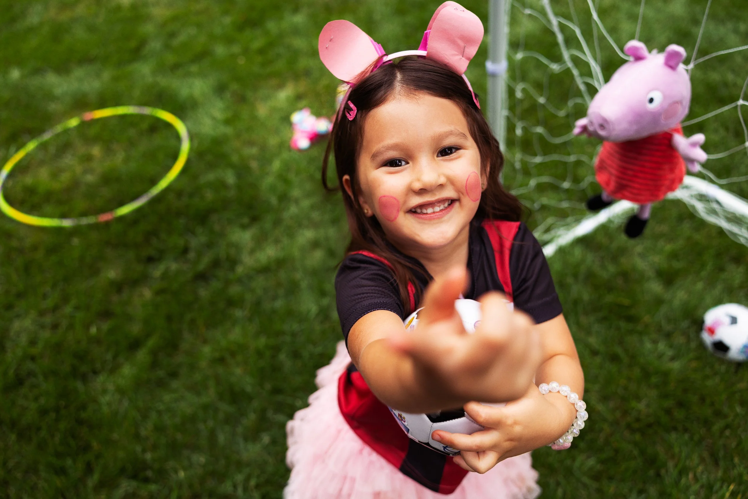 Young girl dressed as Peppa Pig, wearing pink pig ears headband, smiling outdoors, reaching towards the camera.