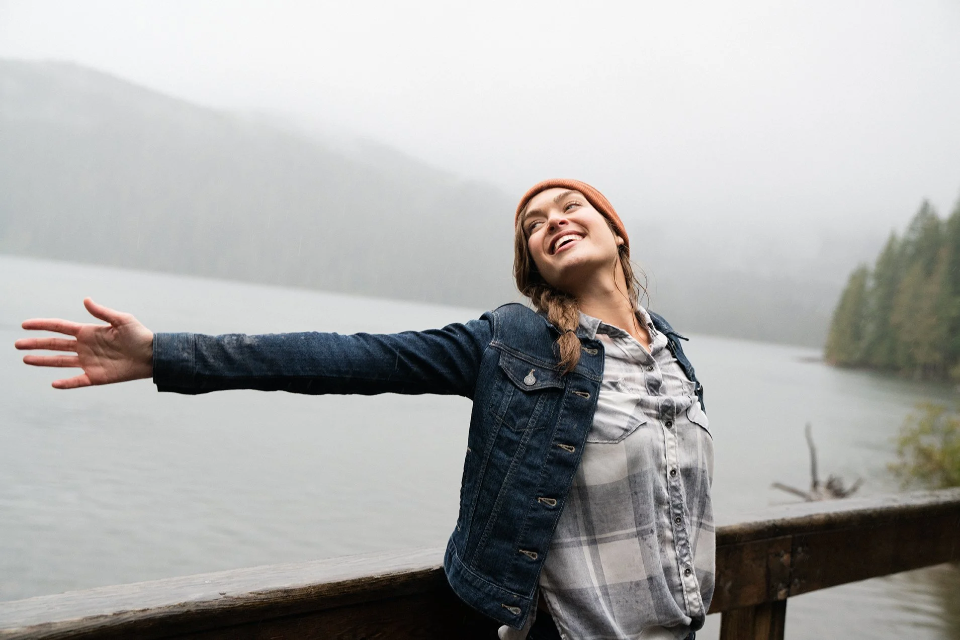 A woman standing on a wooden railing near a lake with mountains in the background, smiling with arms outstretched in a joyful pose, wearing a denim jacket, plaid shirt, and beanie in foggy weather.