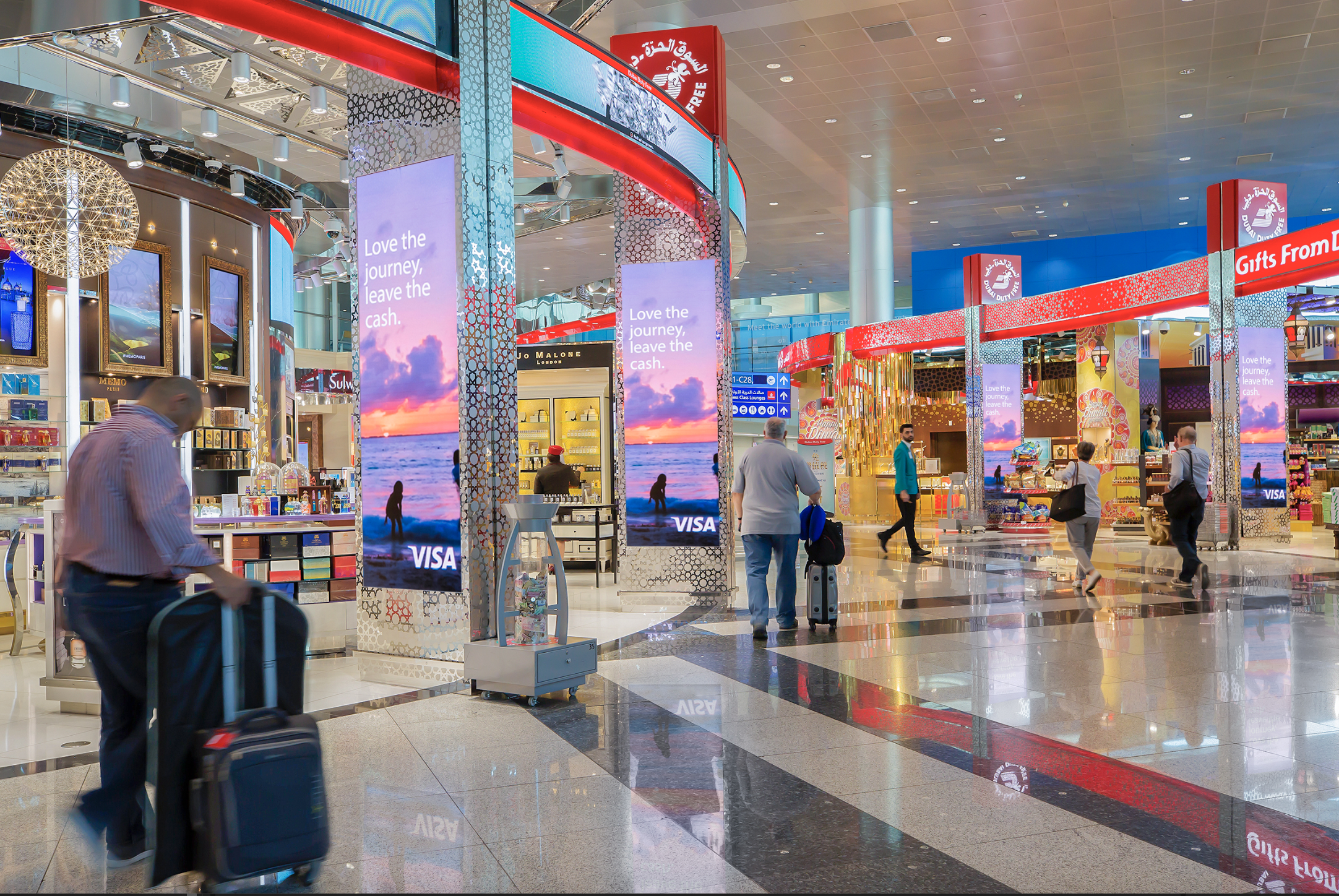 Passengers in an airport terminal shopping and walking past electronic displays and stores, with a colorful ceiling overhead.
