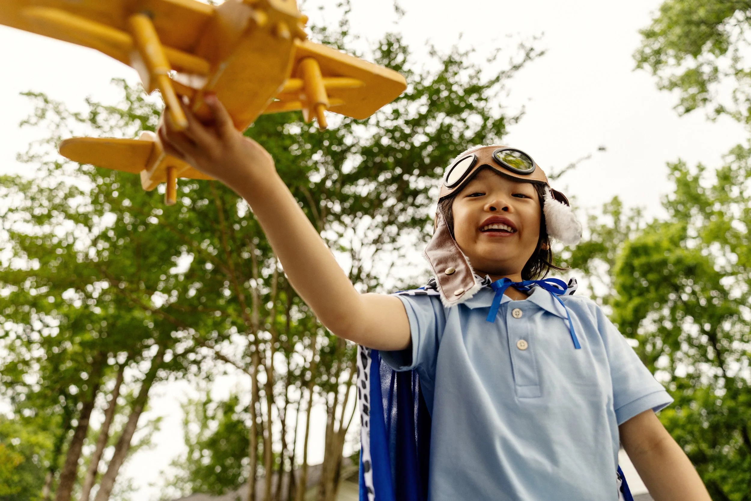 Child dressed as a pilot holding a yellow toy airplane outdoors with trees in the background.
