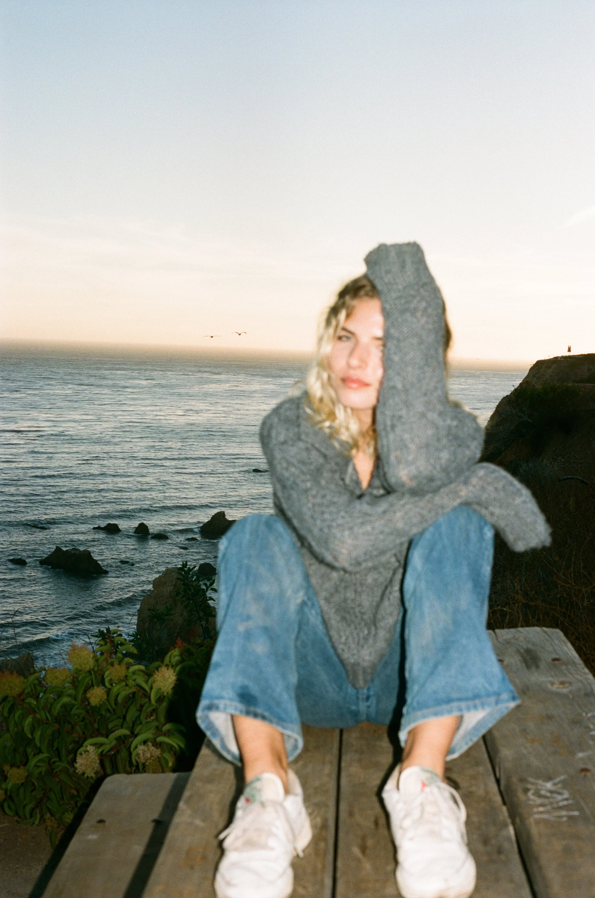 Young woman with wavy blonde hair sitting on a wooden platform near the coast, wearing a gray sweater, blue jeans, and white sneakers, with an ocean and rocky shoreline in the background at sunset.