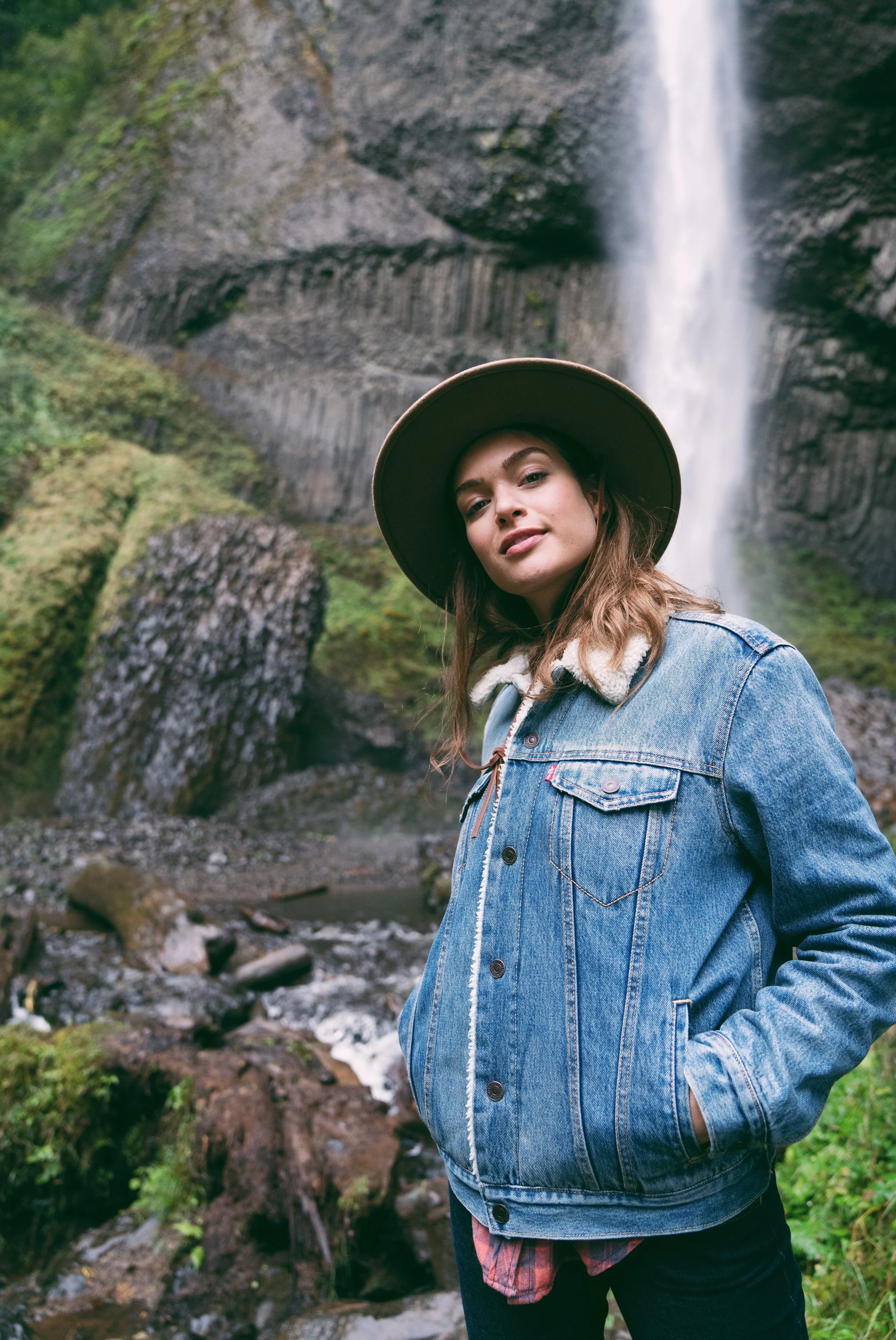 A woman wearing a denim jacket and wide-brimmed hat standing in front of a waterfall in a lush, green forest.