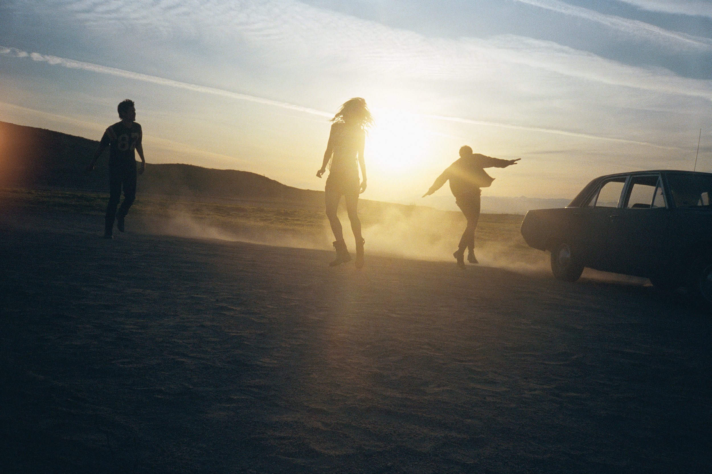 Three people dancing in a desert at sunset, with a dark car parked nearby and dust clouds around their feet.