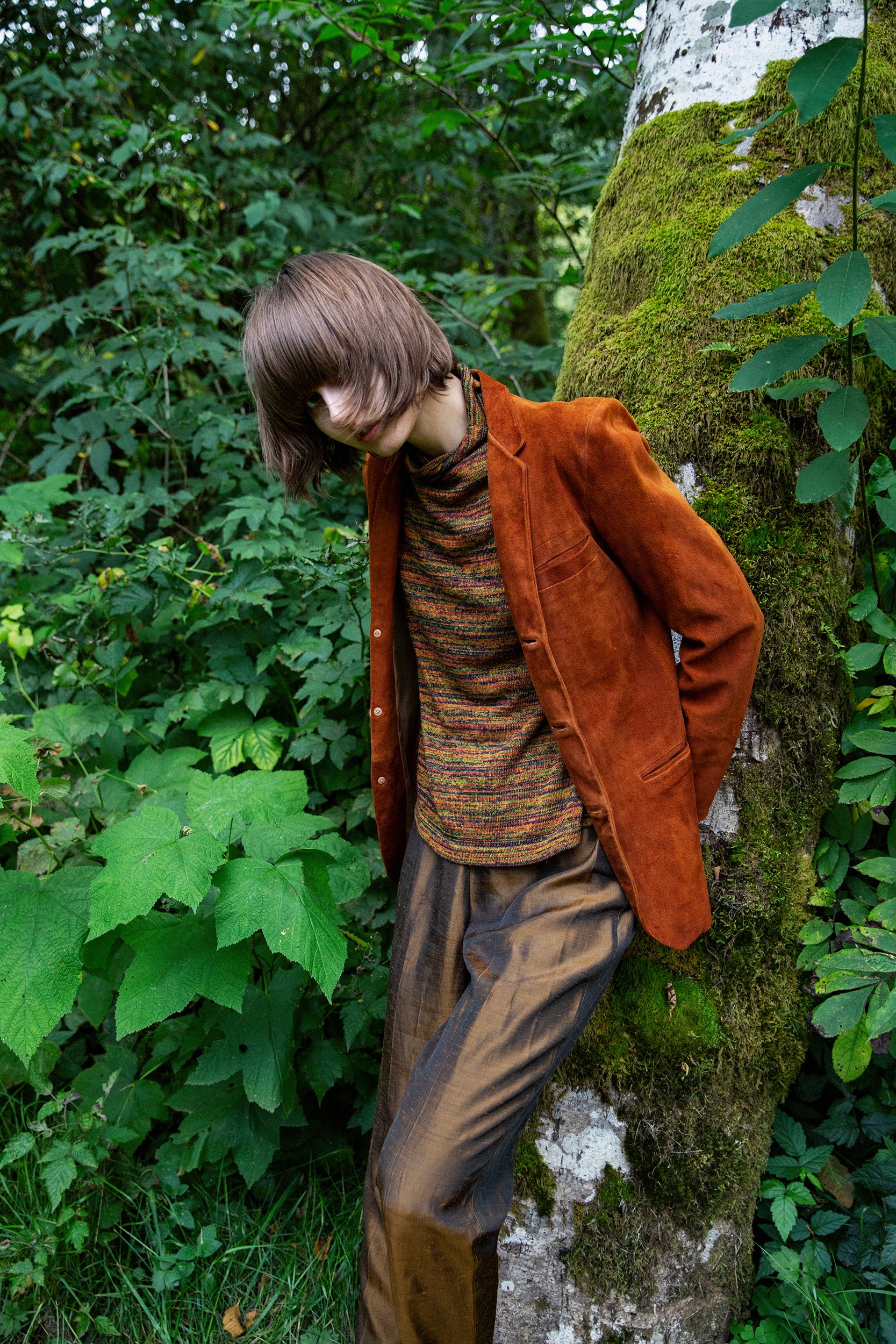 Person leaning against a moss-covered tree in a dense green forest, wearing a brown suede jacket and loose brown trousers.