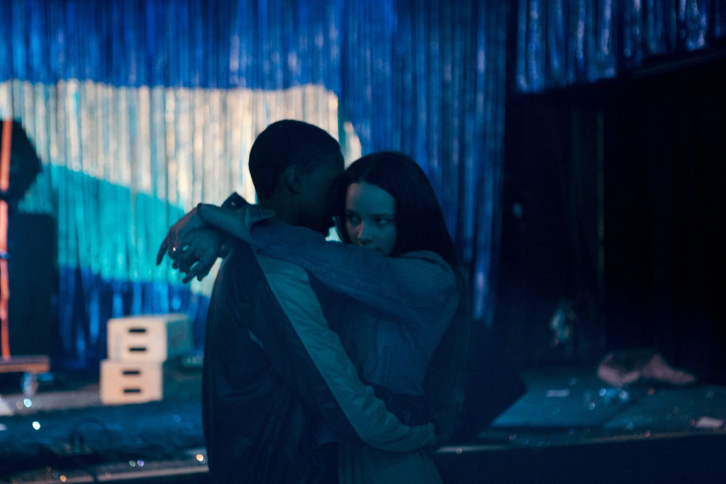 A couple dance closely in a dimly lit, colorful venue with metallic blue curtains, with another person faintly visible in the background.