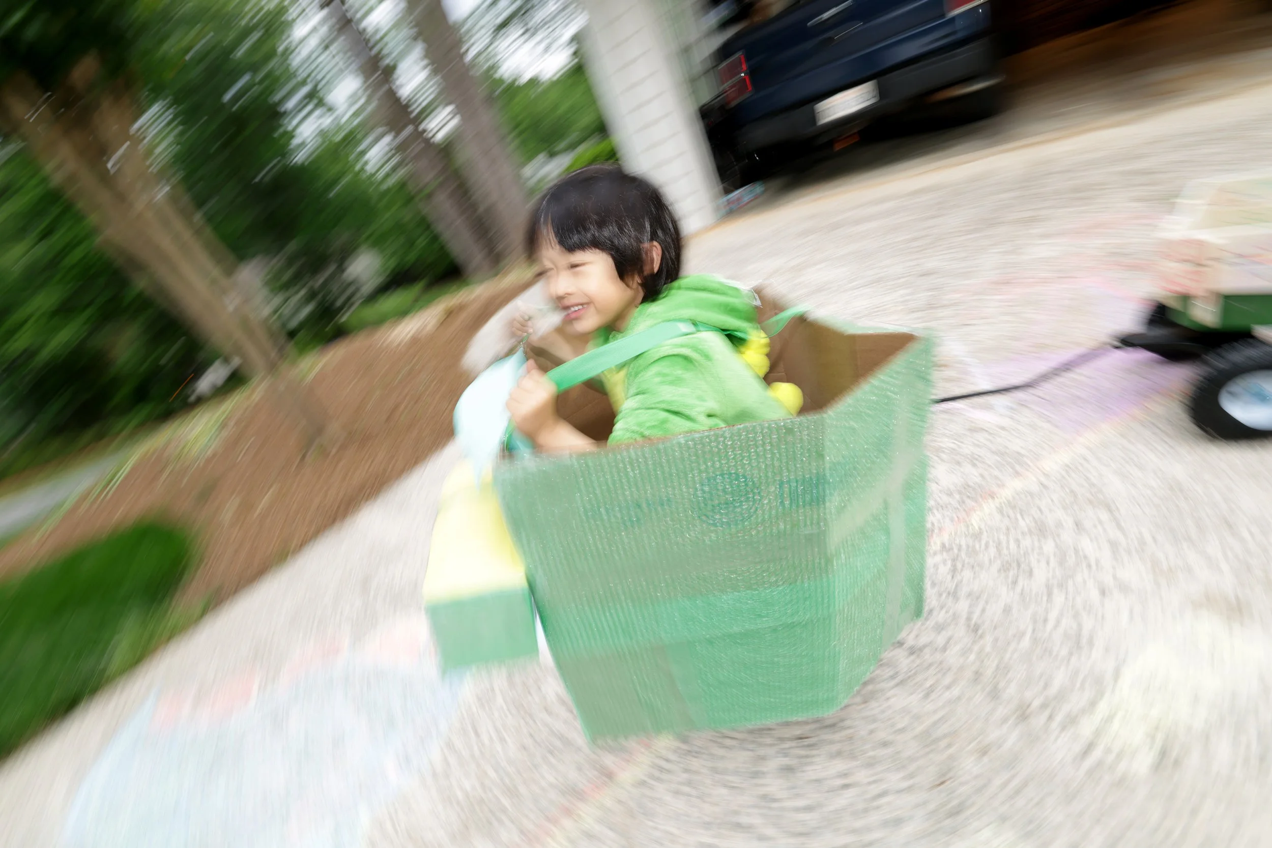Child with dark hair riding in a green stroller or cart outdoors, smiling and dressed in a bright green jacket, with motion blur background of trees, driveway, and a black vehicle.