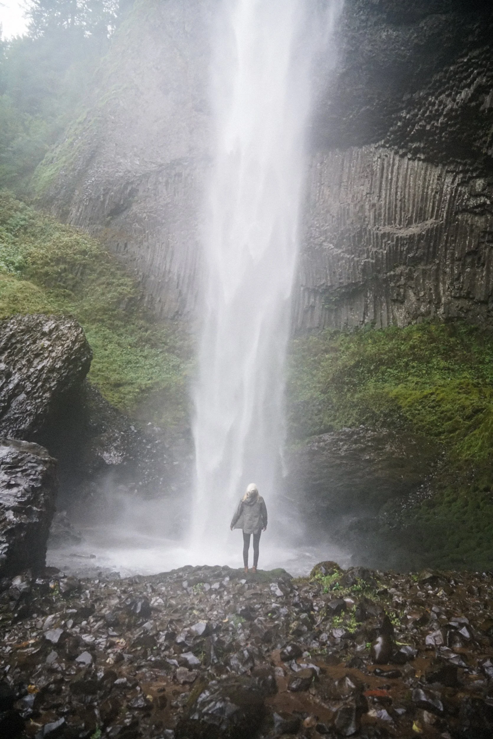 A person standing in front of a tall waterfall surrounded by rocks and green mossy cliffs.