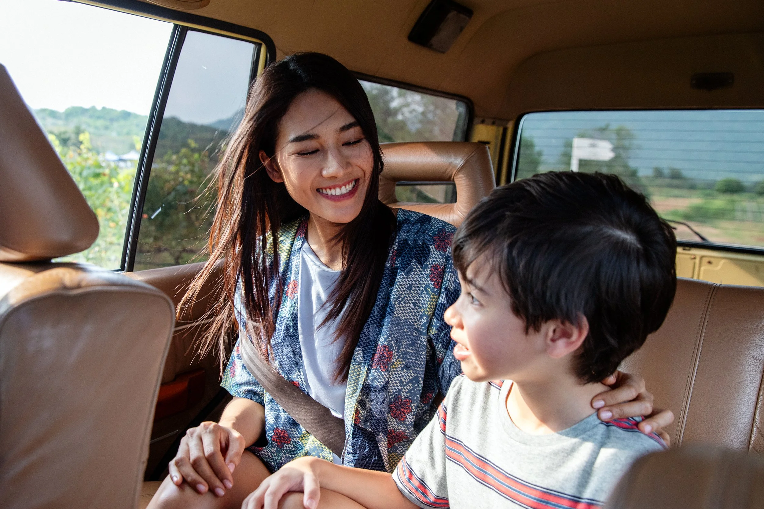 A woman and a young boy sitting in the backseat of a vehicle, smiling at each other during a sunny day.