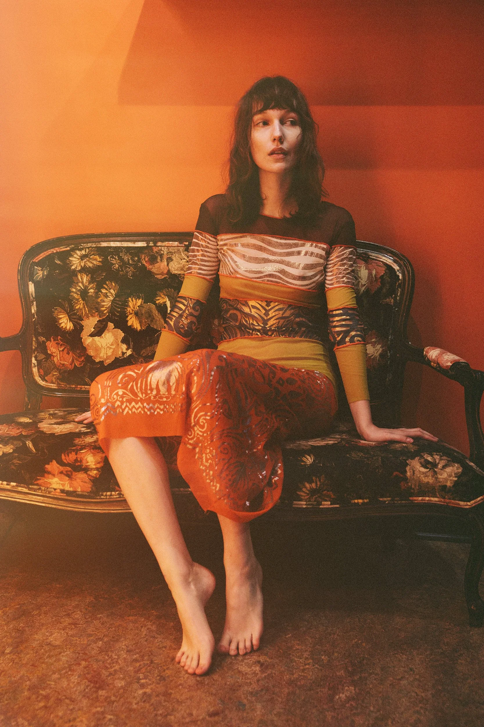 A woman sitting barefoot on a floral upholstered vintage sofa against a warm orange wall, wearing a patterned, multicolored dress with long sleeves.