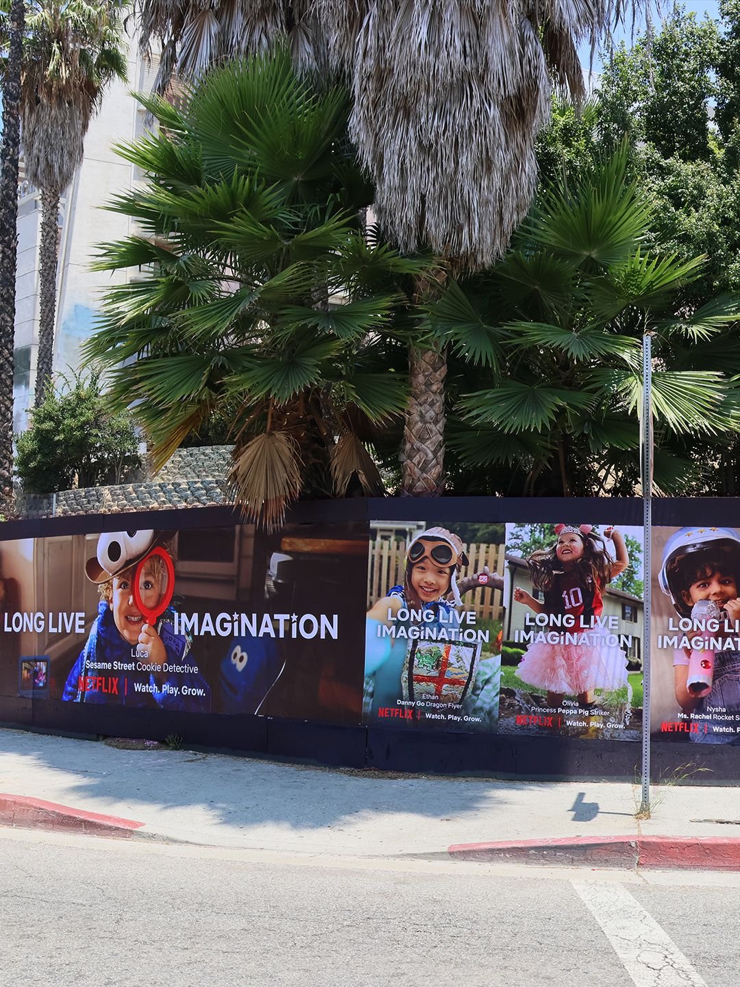 Colorful billboard advertisement featuring four children and promoting Netflix's 'Long Live Imagination' series, with lush palm trees in the background, on a street corner.