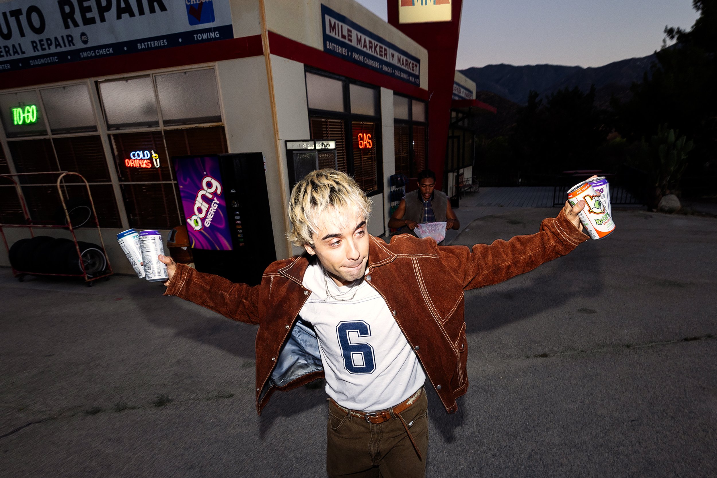 A young man with bleached blonde hair wearing a brown jacket and white t-shirt, holding two cans of Bang energy drinks, standing outdoors in front of an auto repair shop at dusk. There is another person sitting against the building, and various neon 