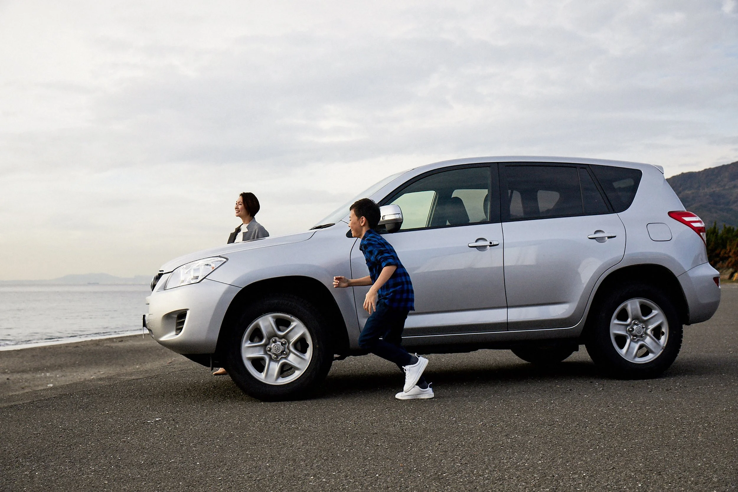 A woman and a boy playing near a silver SUV parked on a coastal road, with the ocean and mountains in the background.