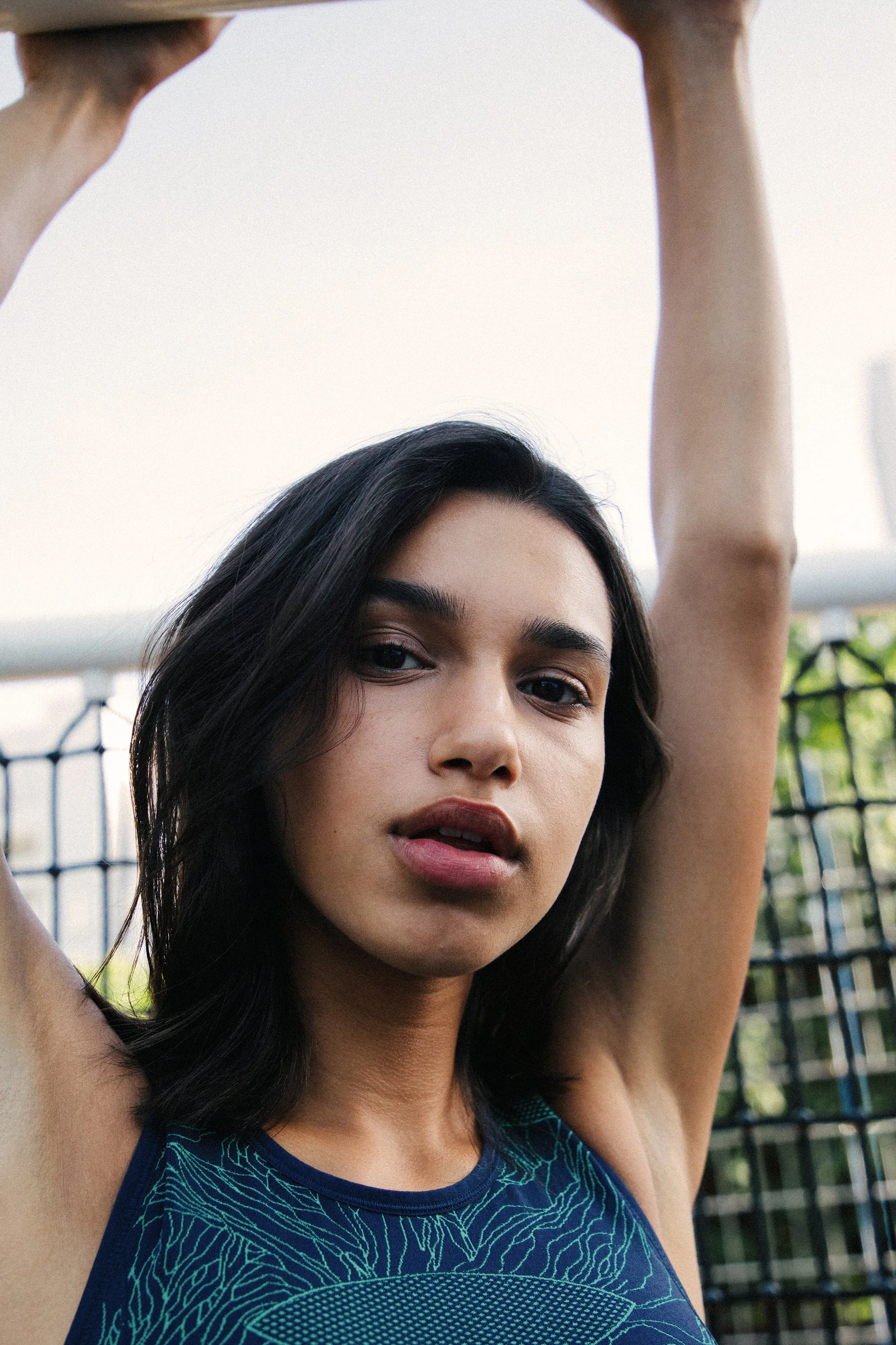 A woman with dark hair and a blue athletic top is raising her arm outdoors, with a fence and greenery in the background.