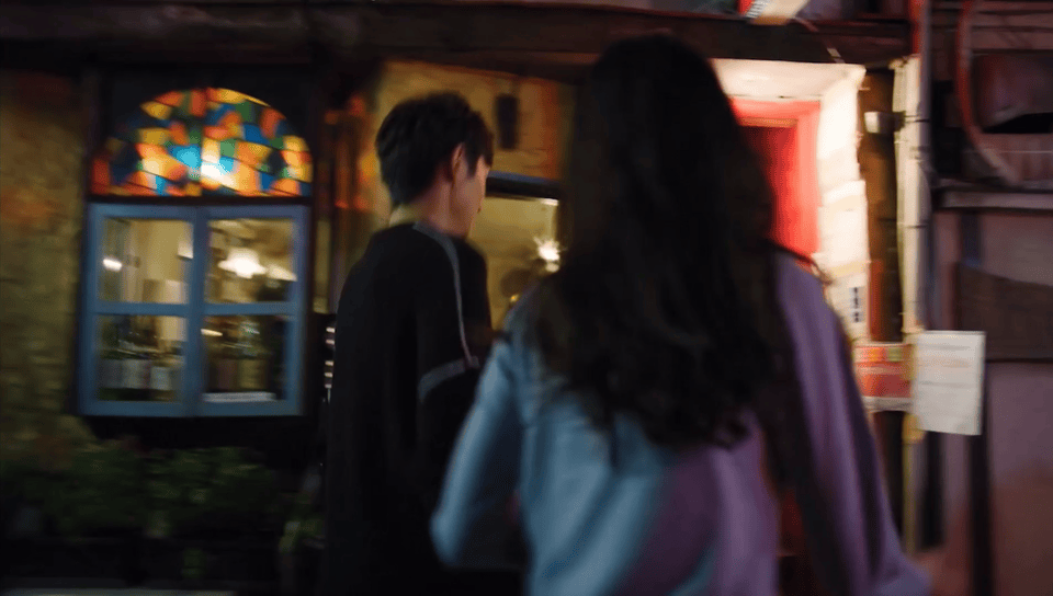 Two people with their back facing the camera walk past an entrance with stained glass window, lit by red lights, in a dimly lit setting.