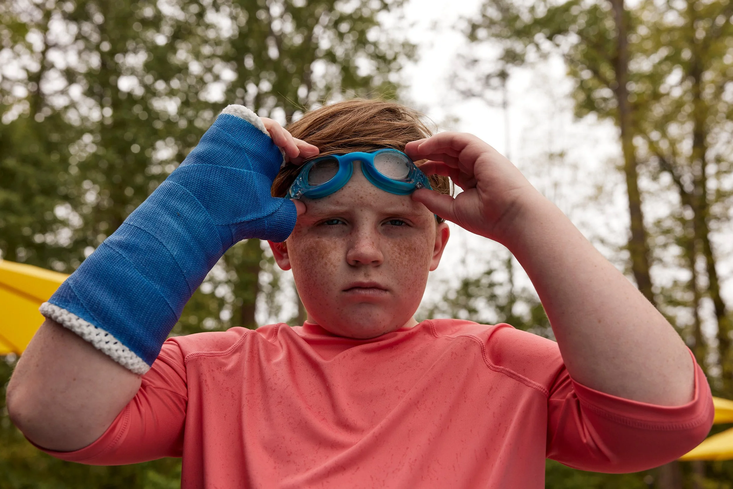 A young boy wearing a coral-colored shirt putting on blue swimming goggles with a serious expression, standing outdoors in a wooded area.