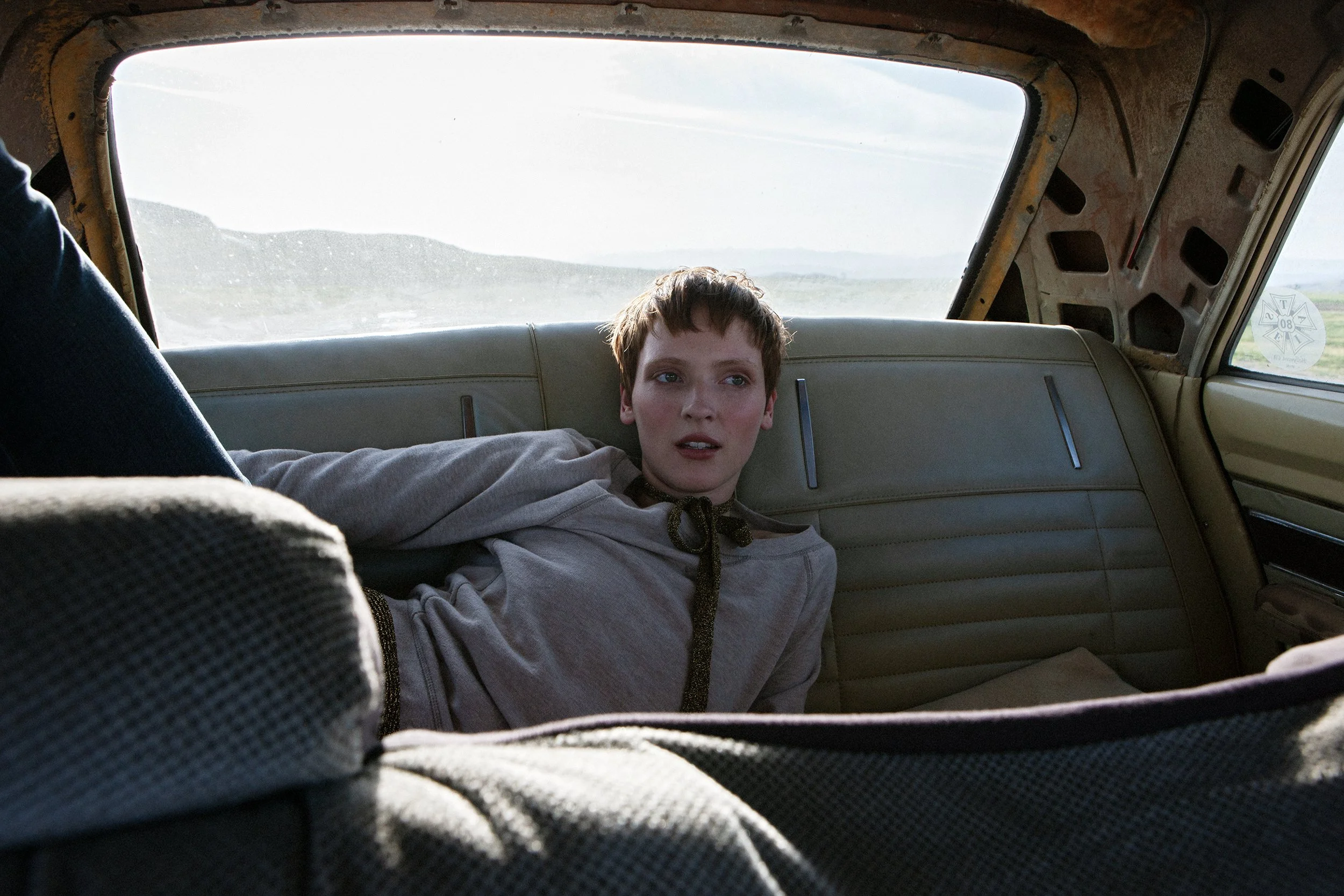 A young boy with short hair wearing a grey sweatshirt lying on the backseat of a vehicle, looking out the window at a rural landscape with mountains in the distance.