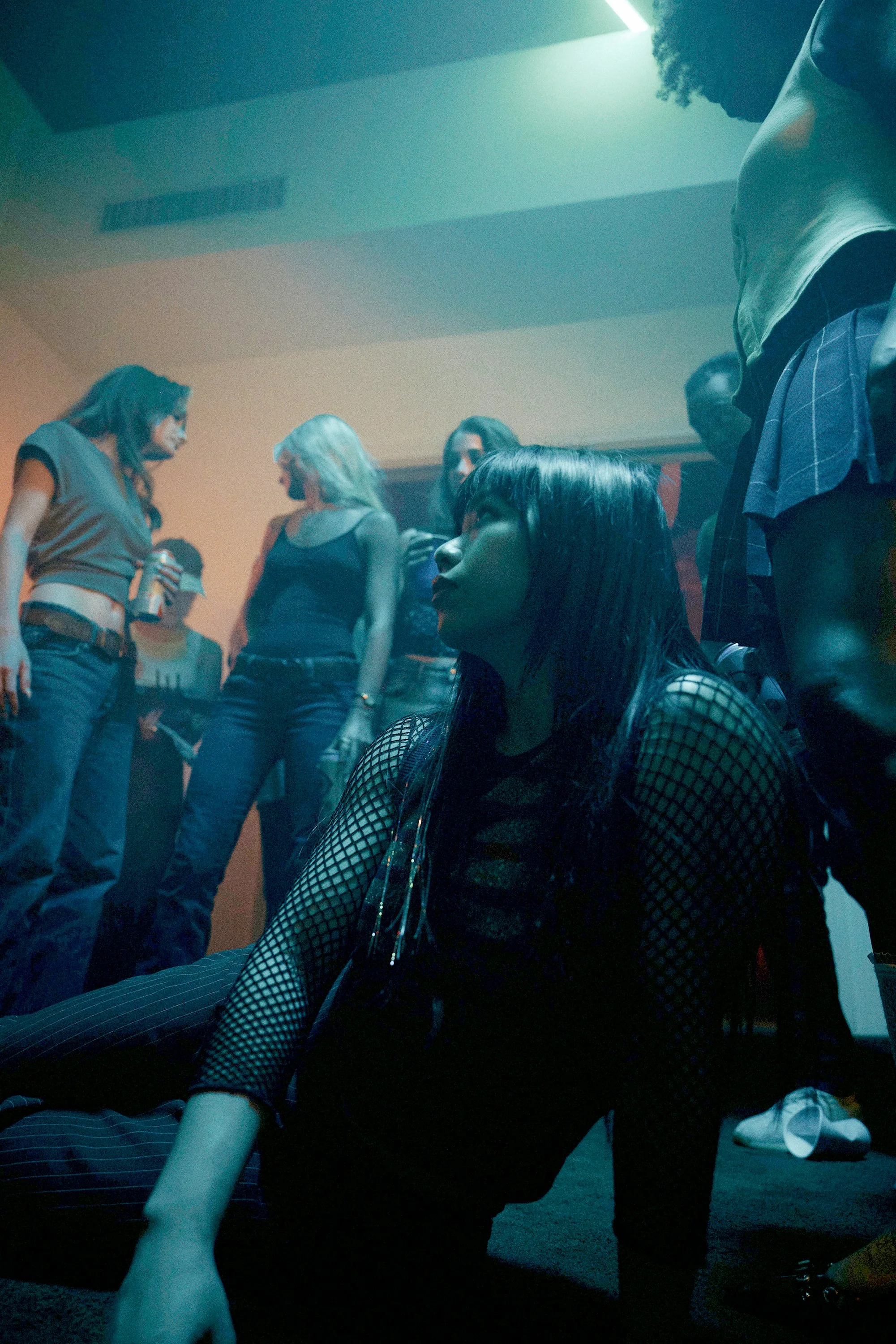 A woman with long dark hair, dressed in fishnet clothing, sitting on the floor in a dimly lit social setting with others standing and talking in the background.