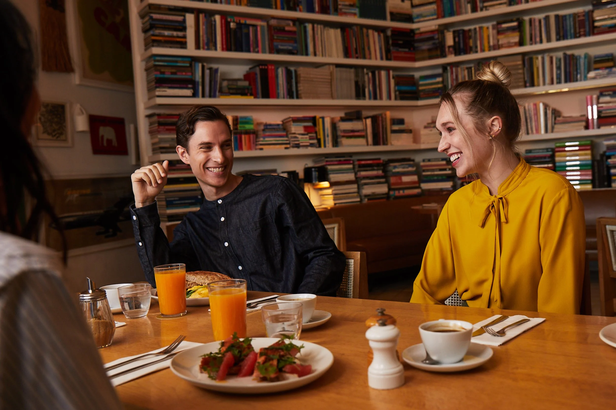 Four people sitting at a restaurant table, smiling and enjoying each other's company. The table has plates of food, glasses of orange juice, and coffee cups, with a background of bookshelves filled with books.