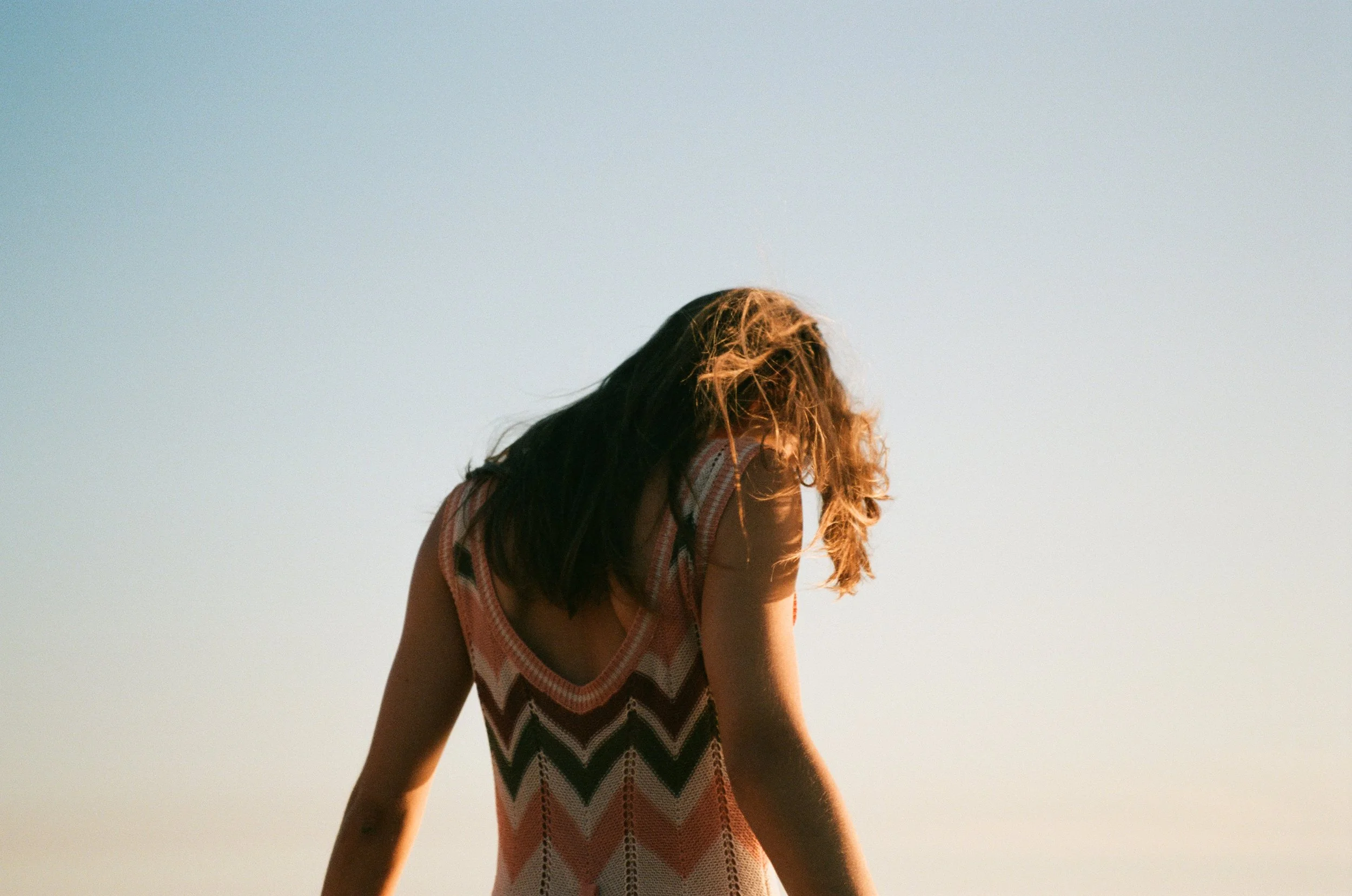 A person with long dark hair in a loose, colorful, zigzag-patterned sleeveless dress stands outdoors against a clear sky, looking downward with their face turned away.