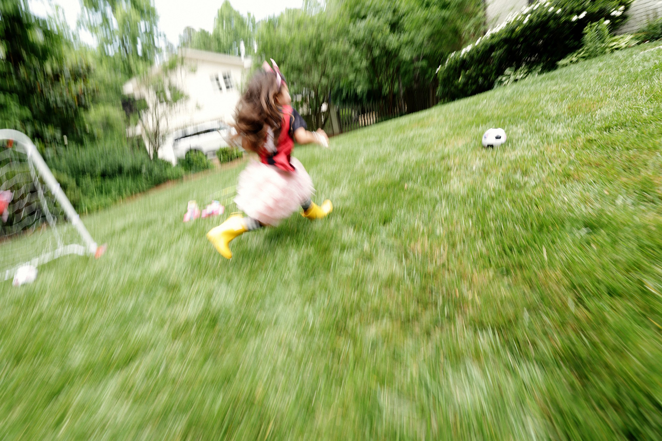 A young girl running outdoors on a grassy lawn with a soccer ball nearby, wearing yellow rain boots, a black top, a pink tutu, and a headband with pink accessories.